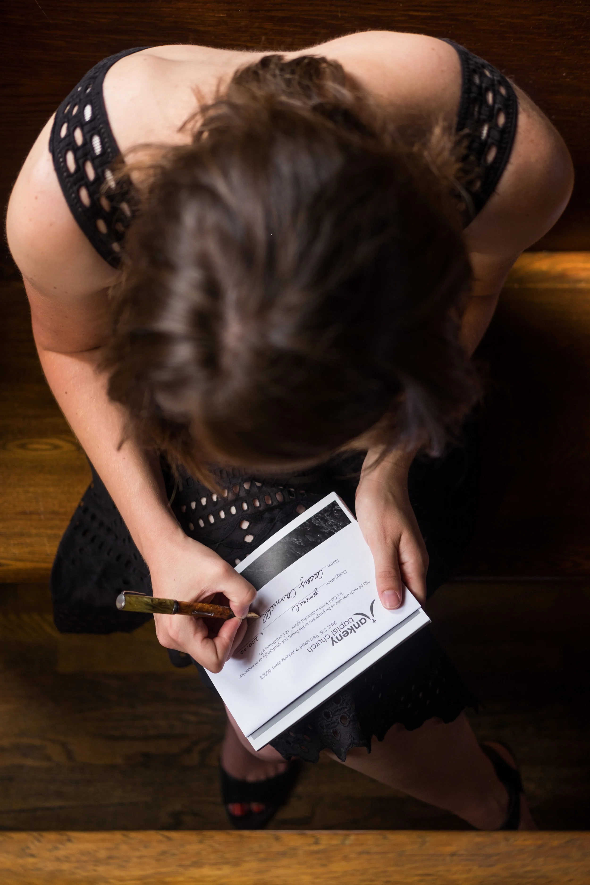 A woman wearing a black dress with cut-out patterns sitting on a wooden bench, signing a program or certificate with a gold-colored pen.