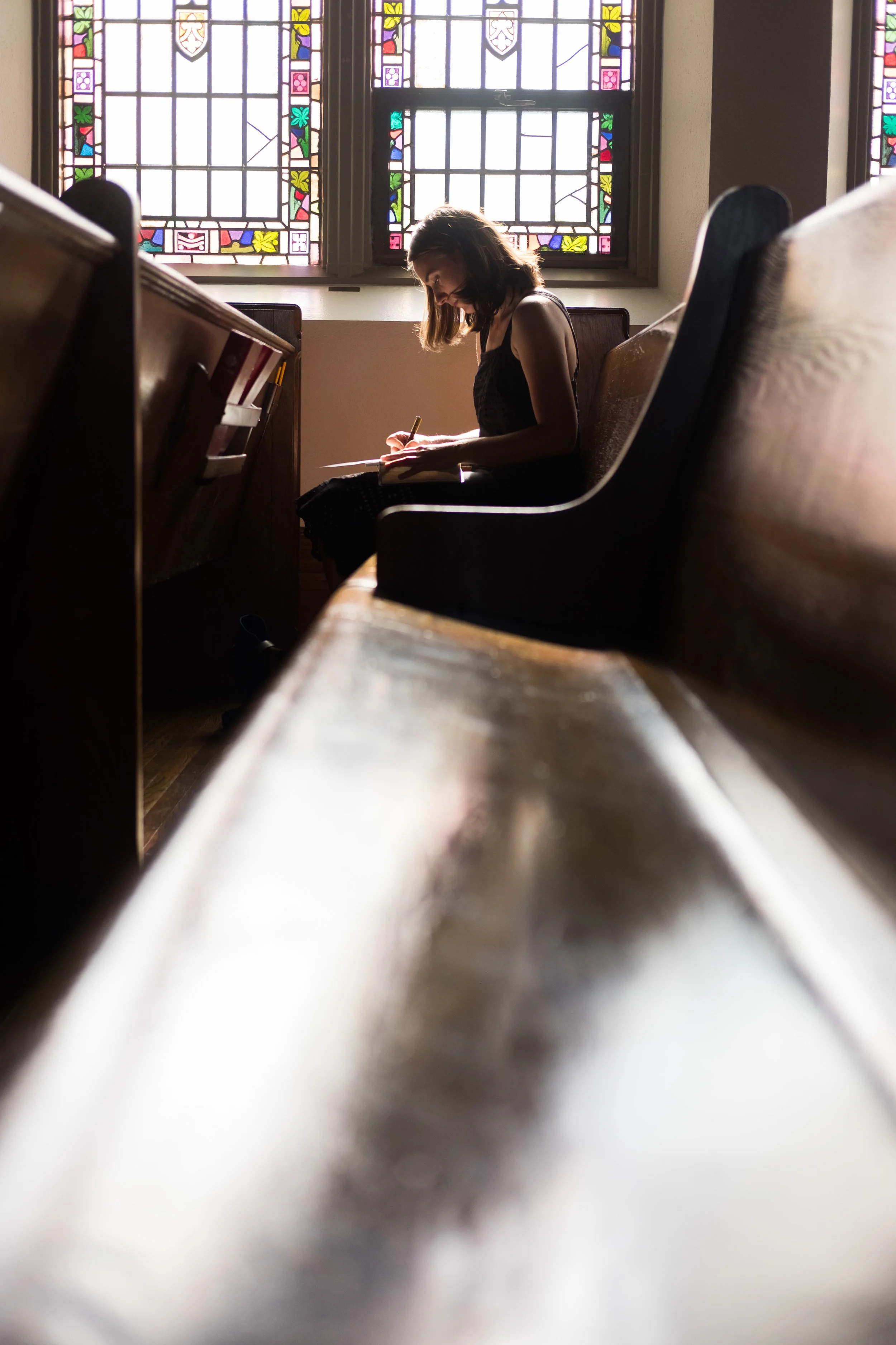 A young woman sitting in a pew inside a church, writing in a notebook, with colorful stained glass windows behind her.