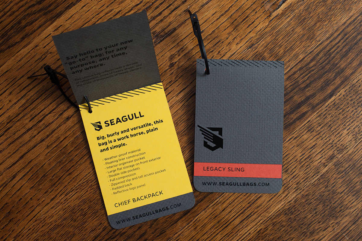 Two luggage tags on a wooden surface, one yellow with black text and the other gray with black and red elements.