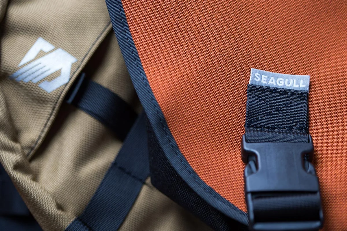 Close-up of two backpacks, one tan with a white logo and one orange with a black clasp and a small label that reads 'SEAGULL'.
