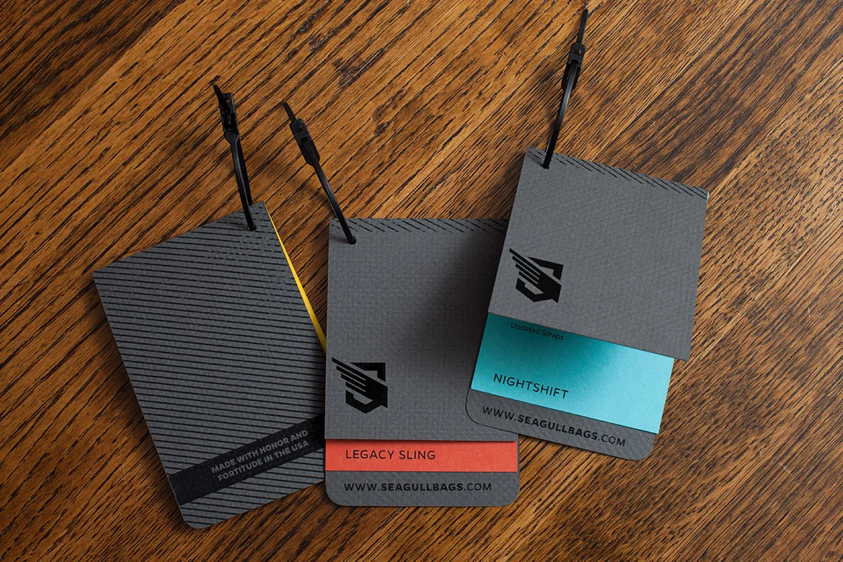 Three tags from Seagull Bags showcasing the 'Legacy Sling' and 'Nightshift' models, attached to a wooden surface.