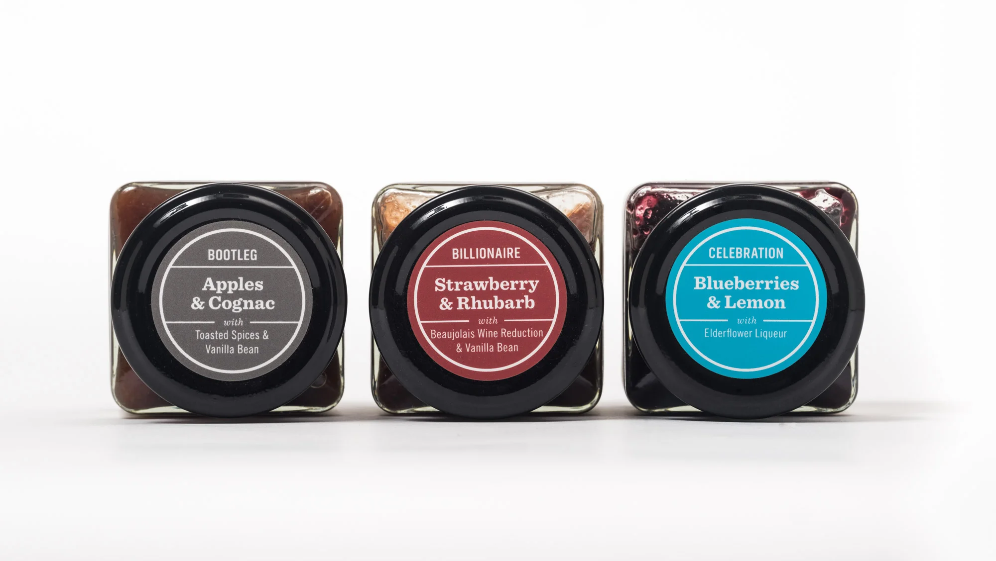Three jars with black lids and colorful labels, from left to right: Apples & Cognac, Strawberry & Rhubarb, Blueberries & Lemon, set against a white background.