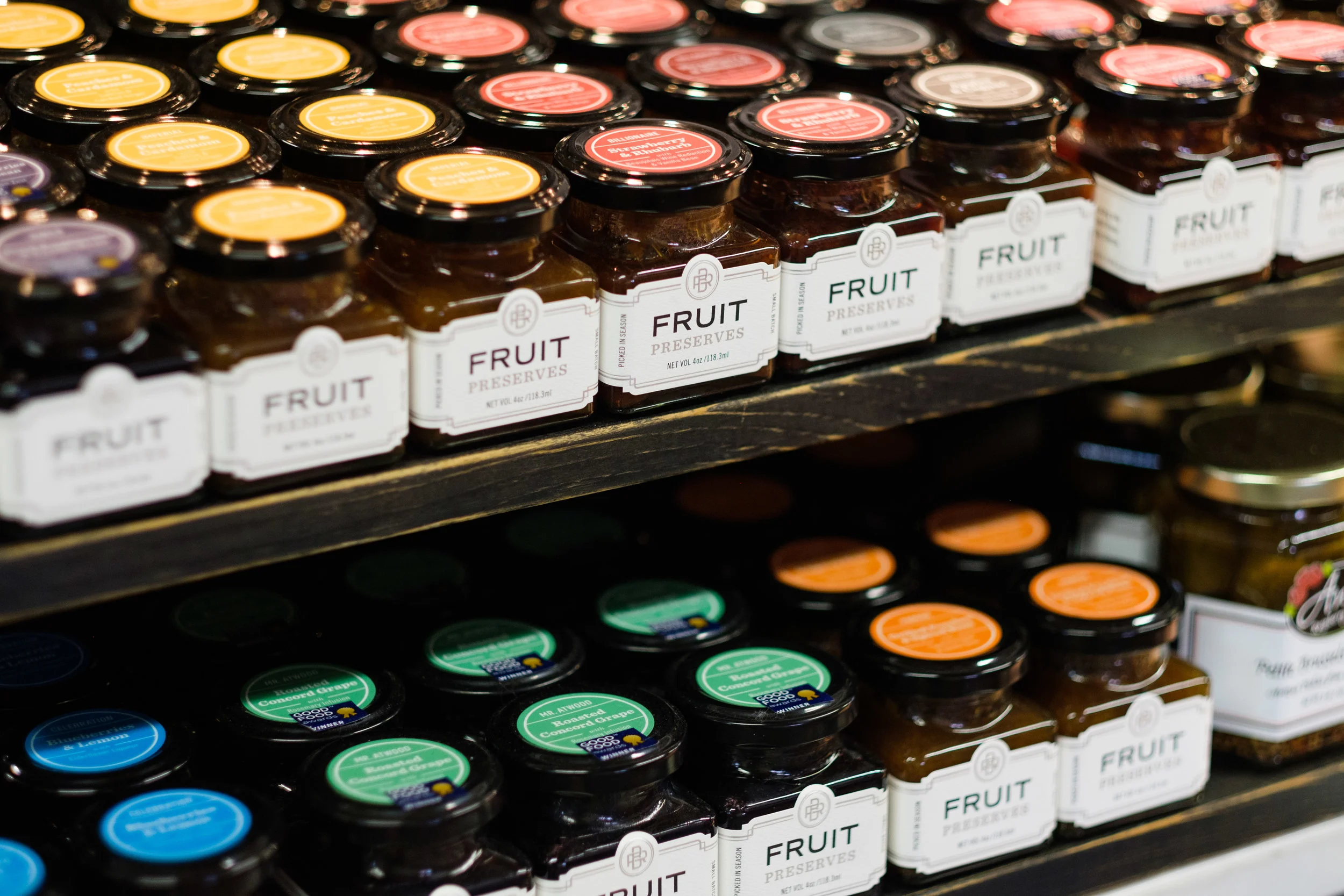 Shelf with jars of fruit preserves in various flavors, including apricot, concord grape, and mixed fruit, with colorful labels and black lids.