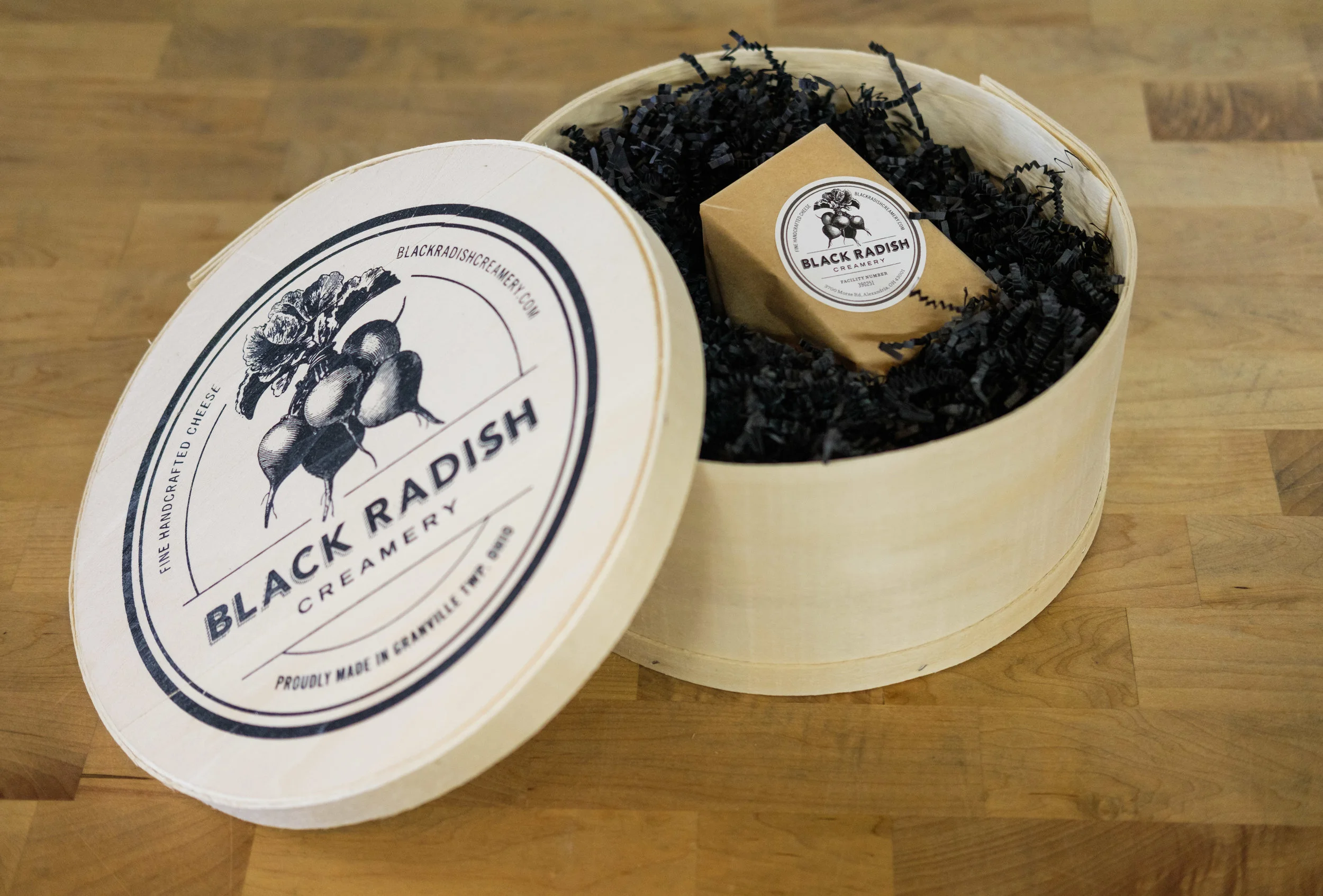 A round wooden box with a lid containing a bar of cheese wrapped in paper inside black shredded paper. The lid is open, revealing the cheese and a label. The label on both the box and the cheese wrapper reads "Black Radish Creamery" with a logo of ra