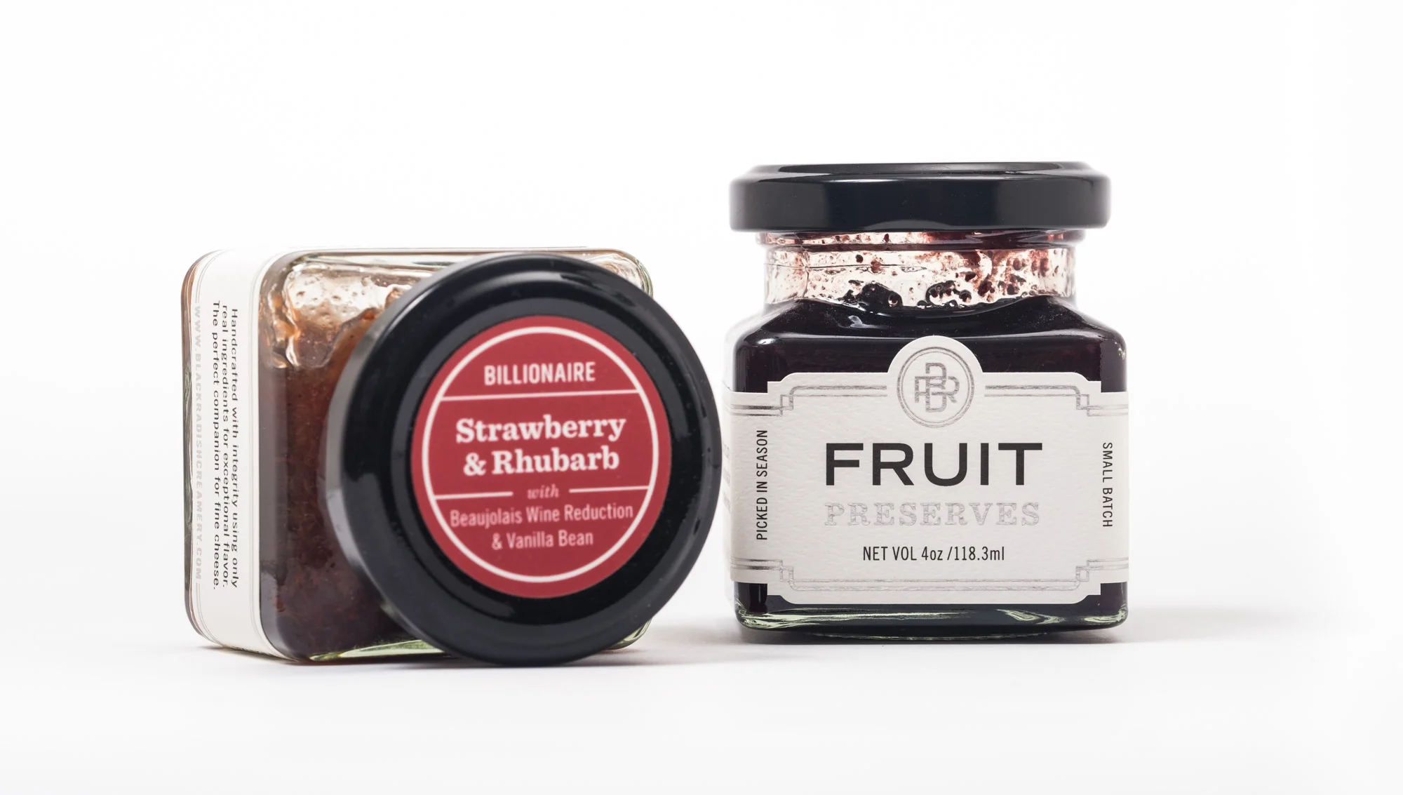 Two jars of fruit preserves, one labeled "Strawberry & Rhubarb" with wine reduction and vanilla bean, and the other labeled "Fruit Preserves" with a dark jelly inside.
