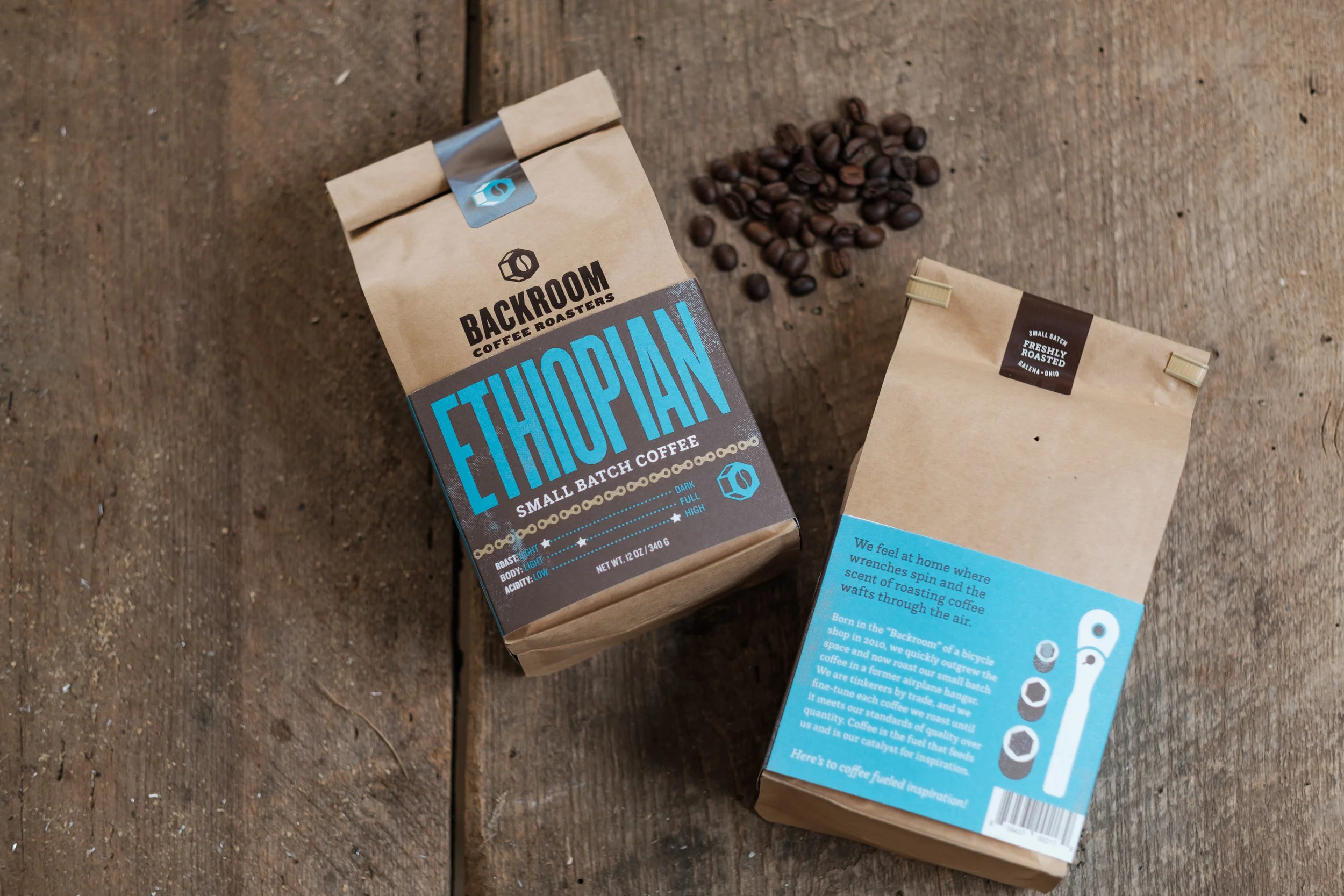 Two brown paper bags of coffee, one labeled 'Ethiopian' from Backroom Coffee Roasters and the other with a blue label and a graphic of coffee pods, placed on a wooden surface with scattered coffee beans.