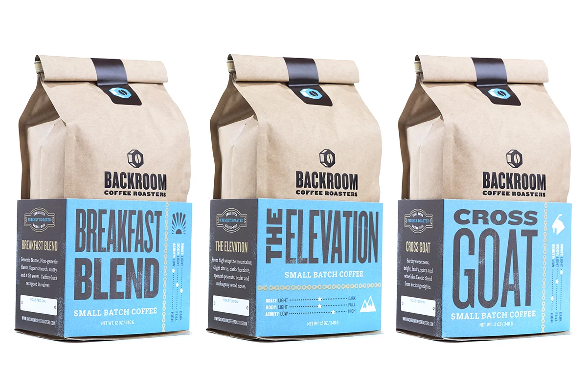 Three small brown paper bags of Backroom Coffee Roasters' small batch coffee, with blue labels indicating different blends: Breakfast Blend, The Elevation, and Cross Goat.