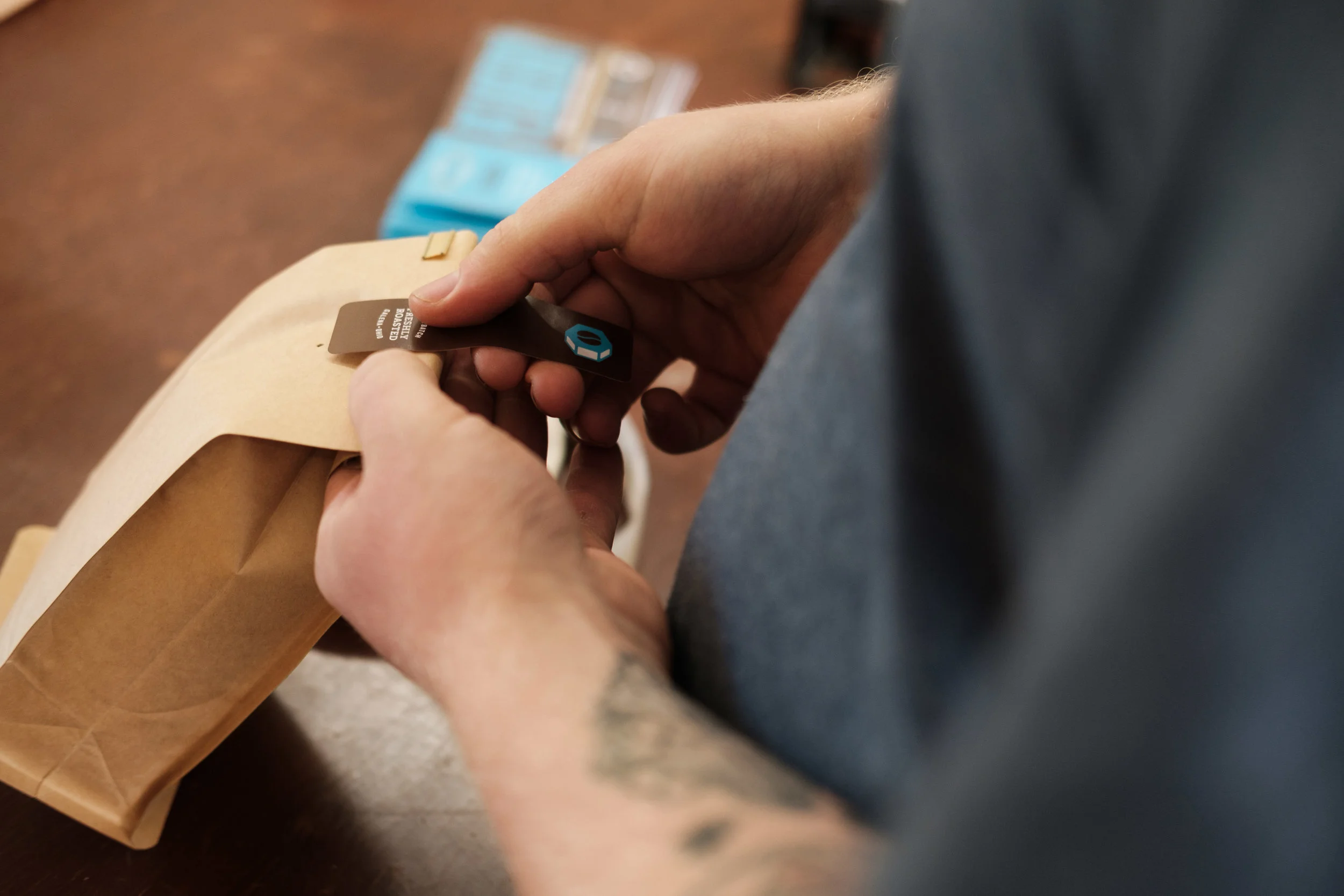 A person holding a brown luggage tag with a black strap, attaching it to a beige paper bag. A blue snack package is visible in the background.