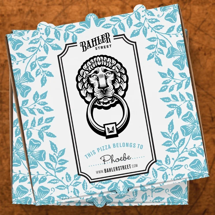 Pizza box with a blue floral and leaf pattern, featuring a lion's head emblem with a ring in its mouth, and text indicating it belongs to Phoebe from Bahler Street.