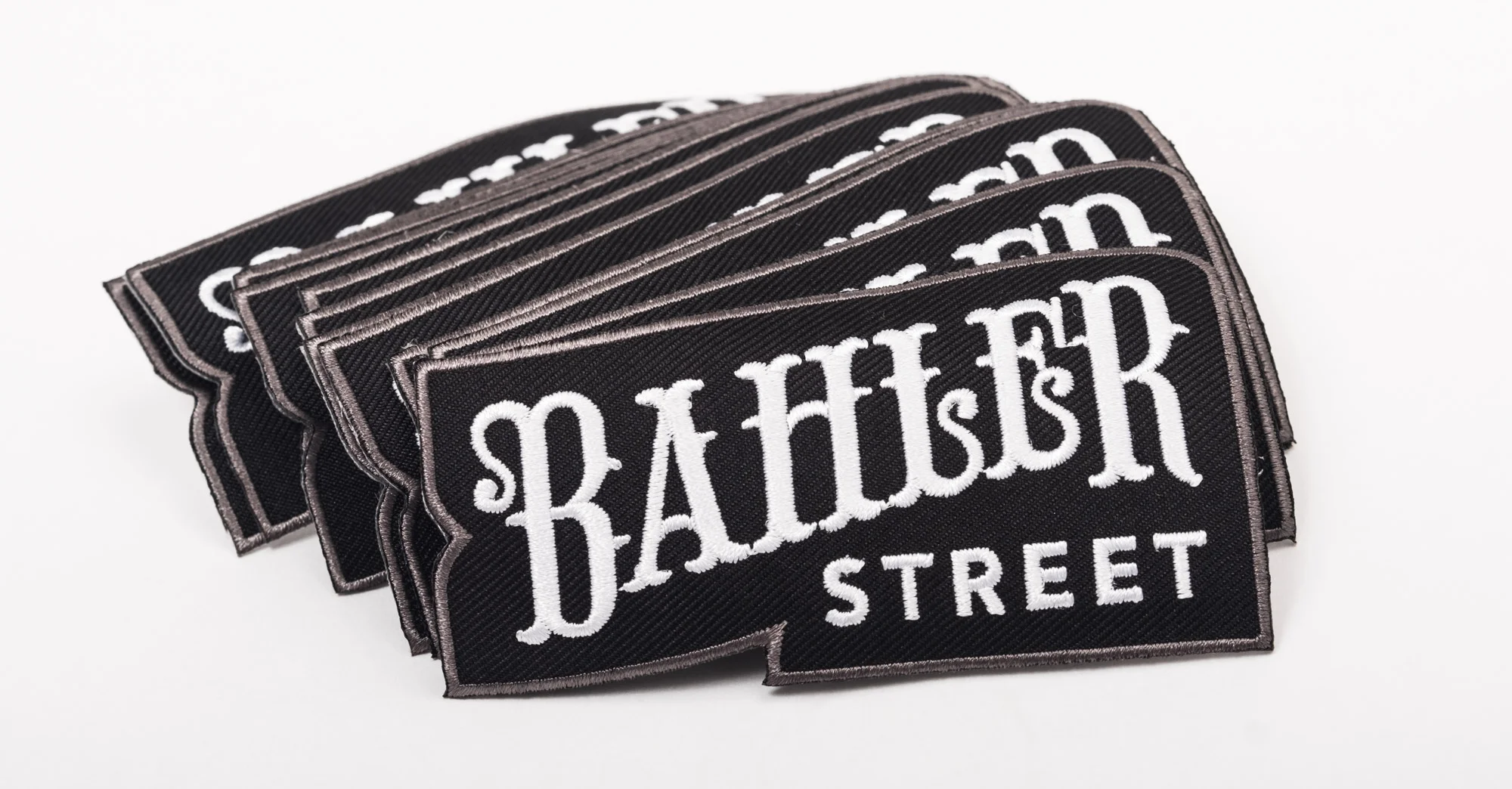 A stack of black fabric patches with white embroidered text reading 'Baldwin Street', edged with a gray border, placed on a white surface.