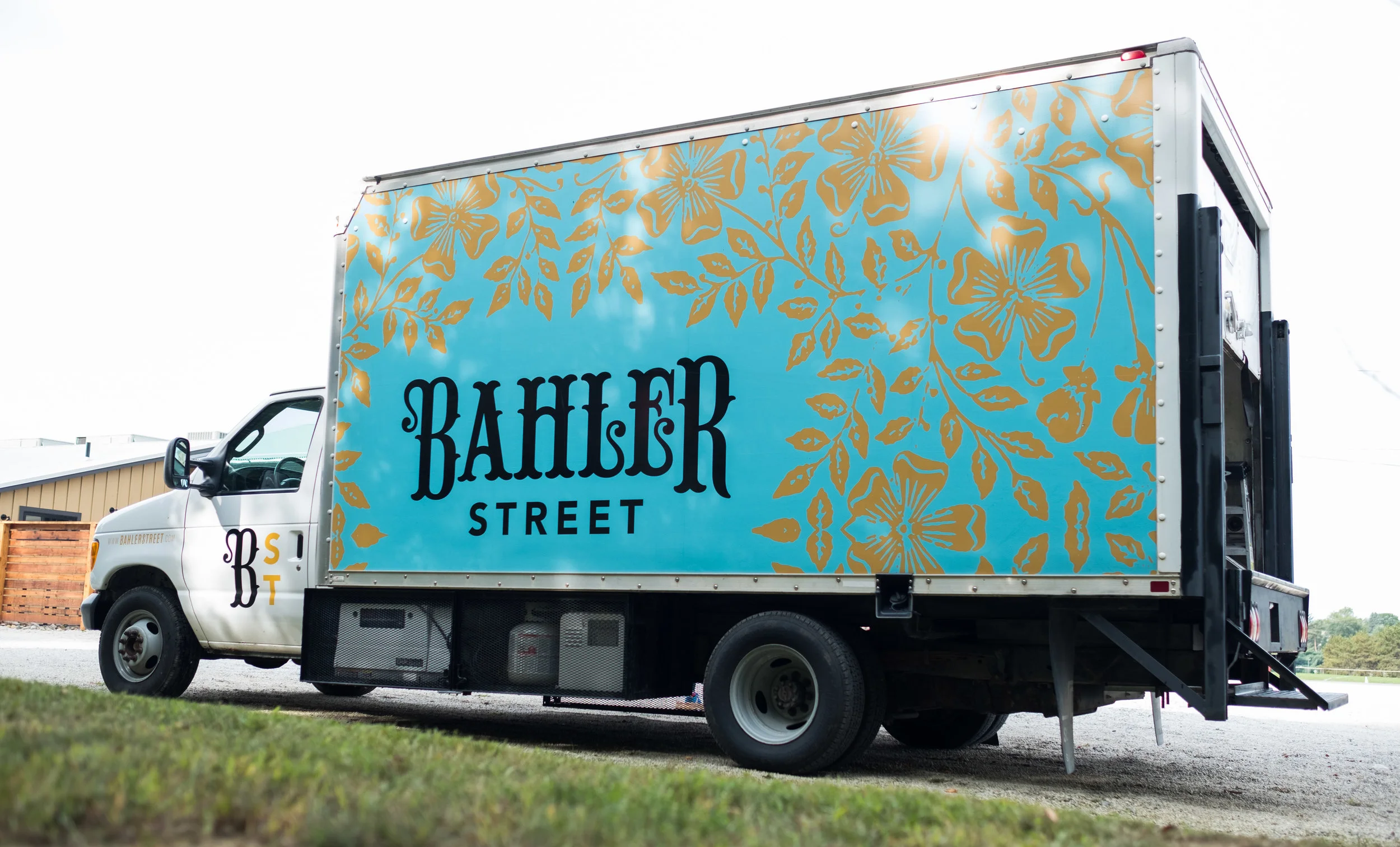 Delivery truck with a bright blue side panel decorated with gold floral patterns and the text 'BAHLER STREET' in black, parked outdoors on grass with a building in the background.