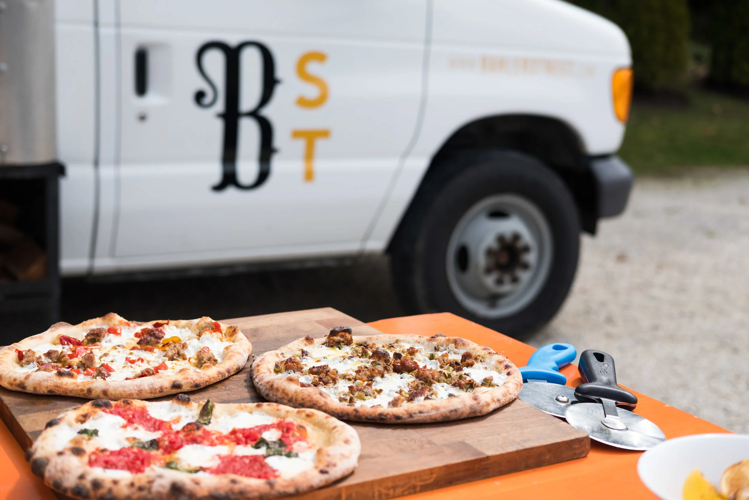 Three pizzas on a wooden board near a food truck, with scissors and a pizza cutter on an orange table.
