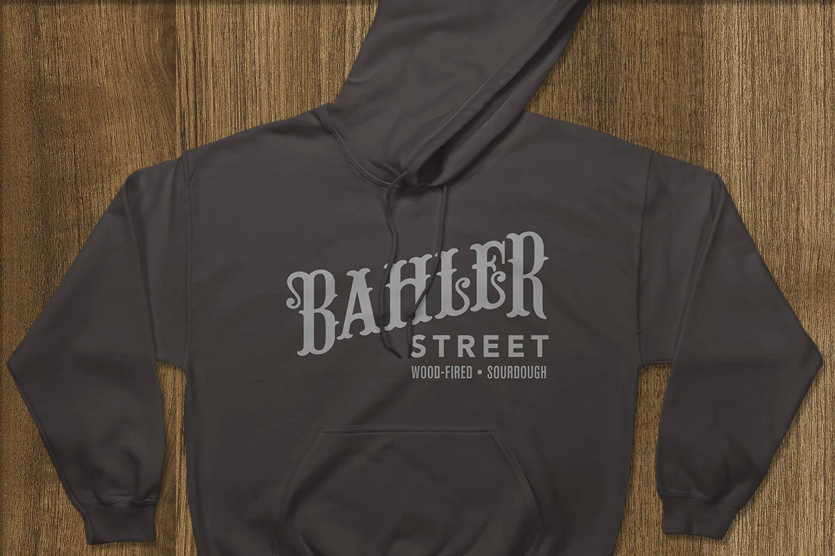 Black hoodie with gray text that reads 'Bamberg Street Wood-Fired Sourdough,' laid flat on a wooden surface.