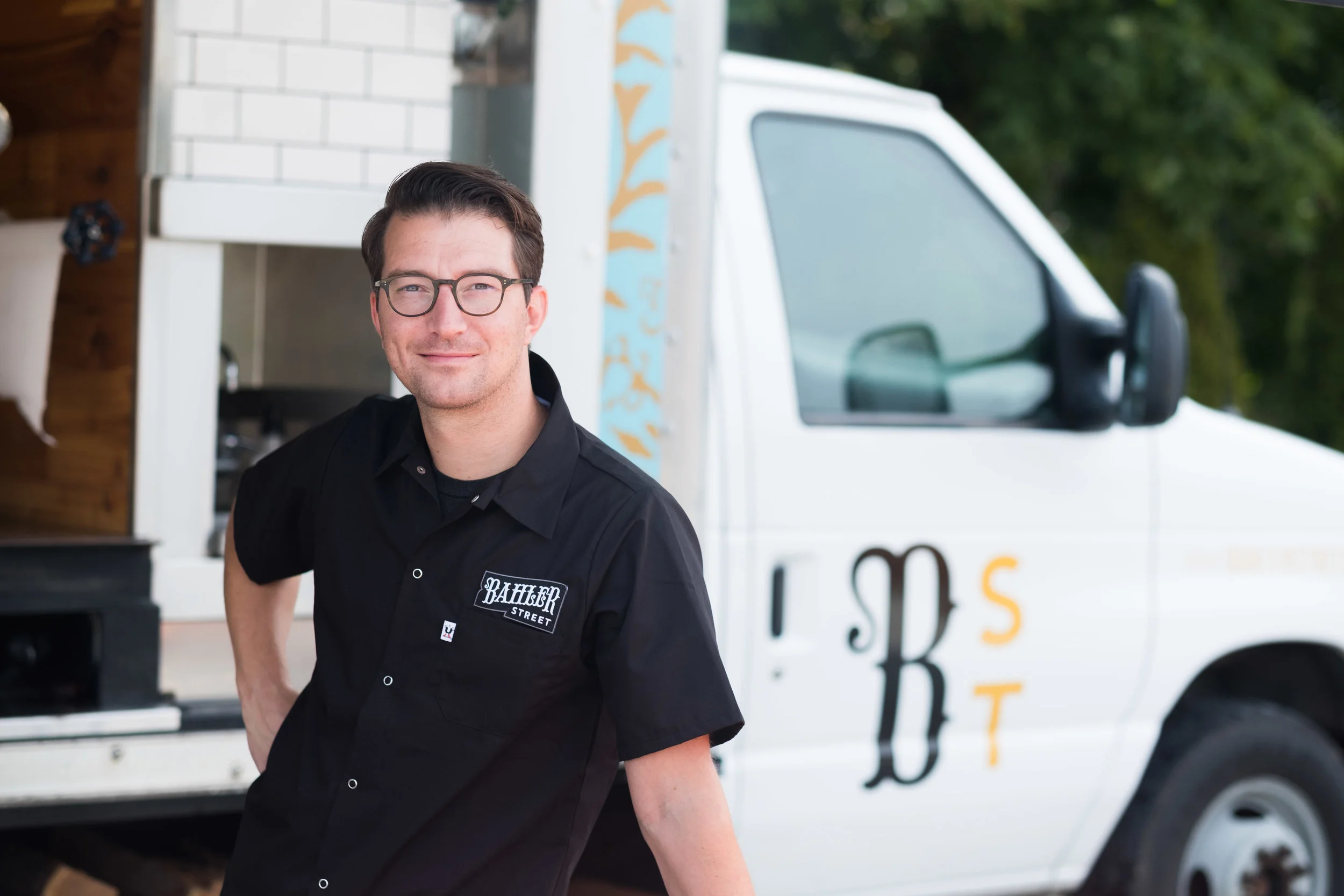 A man with glasses and dark hair, wearing a black shirt with a 'Hambr Street' patch, stands in front of a white food truck labeled 'RSBT'.