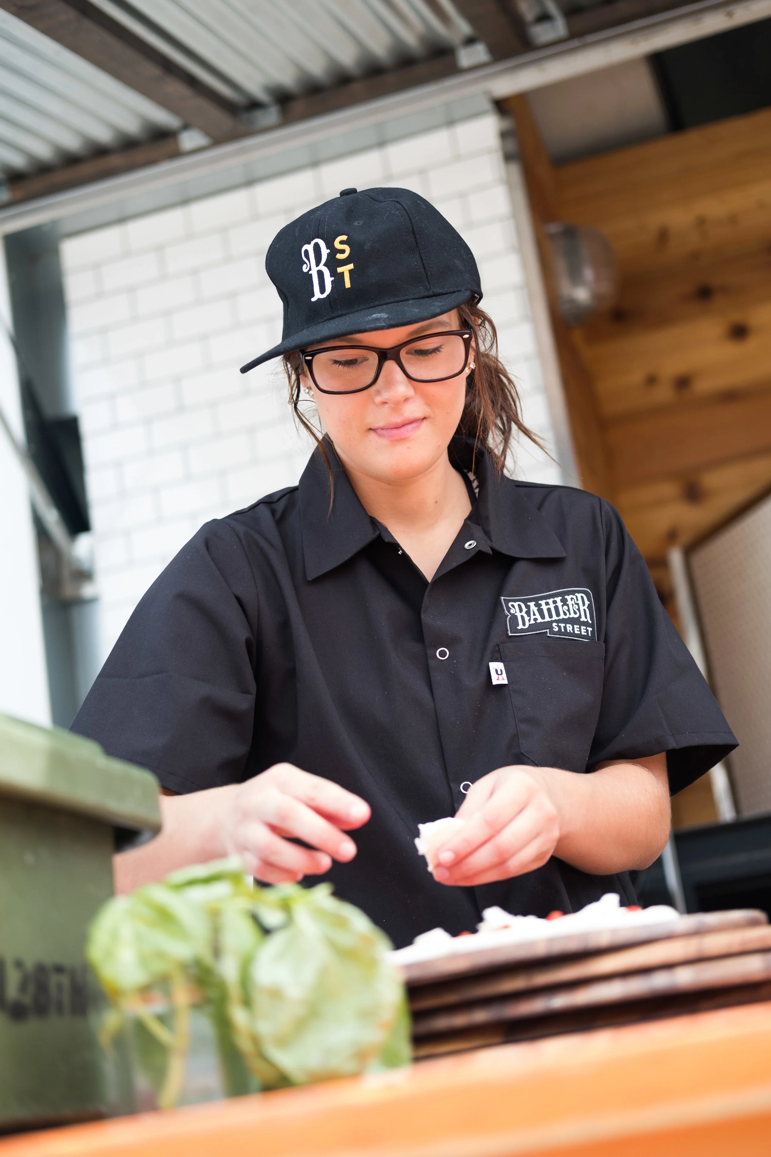 A young woman wearing glasses, a black cap, and a black shirt with a patch that says "Ballers Street" is preparing food at a counter, possibly making a sandwich or similar snack.