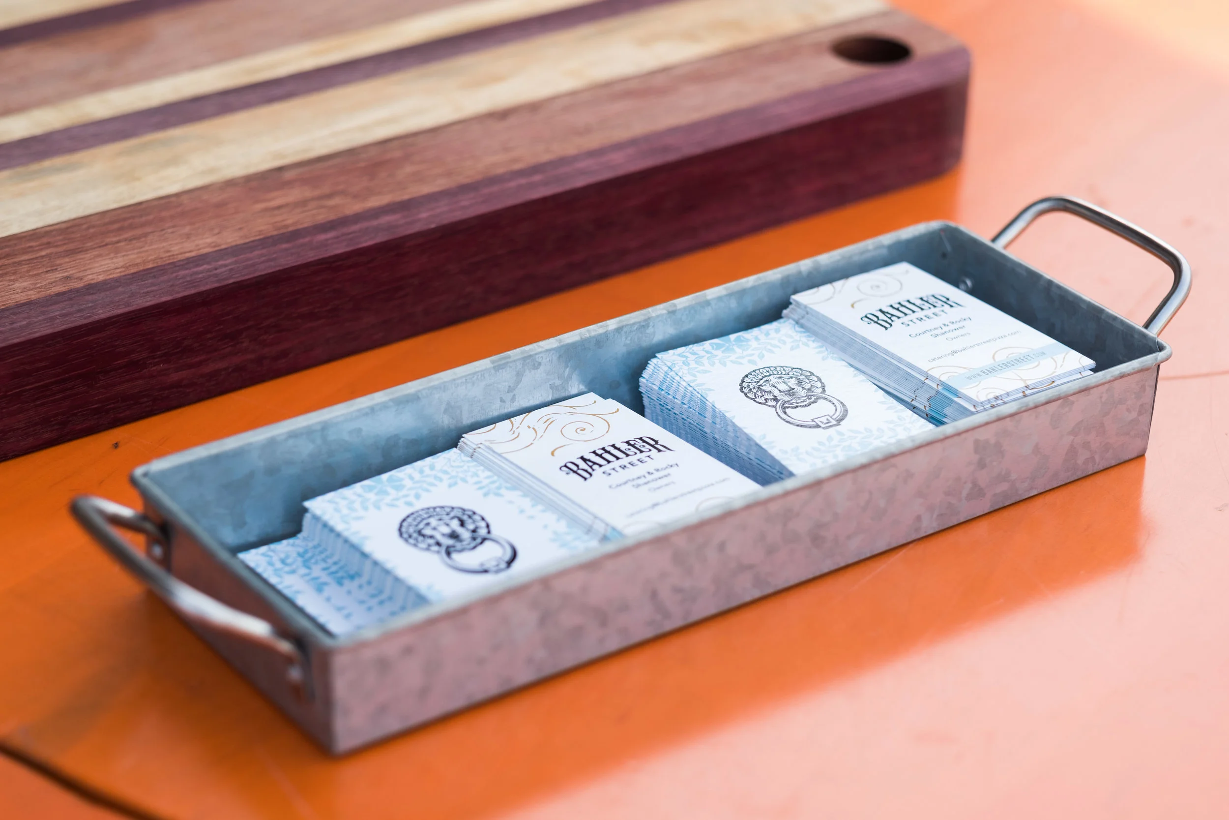 Stacks of business cards inside a metal tray on an orange table with a wooden cutting board in the background.