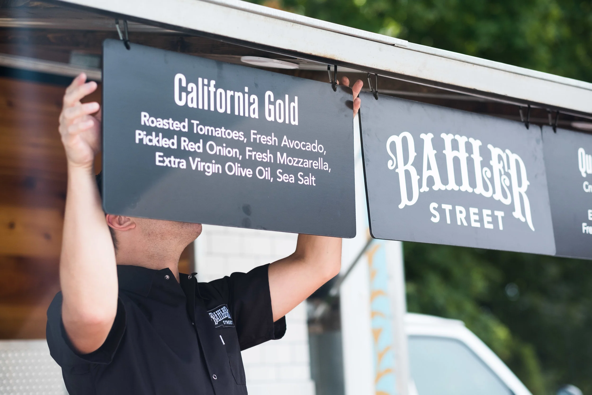 A person is lifting a sign that reads 'California Gold' with a menu of roasted tomatoes, fresh avocado, pickled red onion, fresh mozzarella, extra virgin olive oil, and sea salt, on a street food stand.