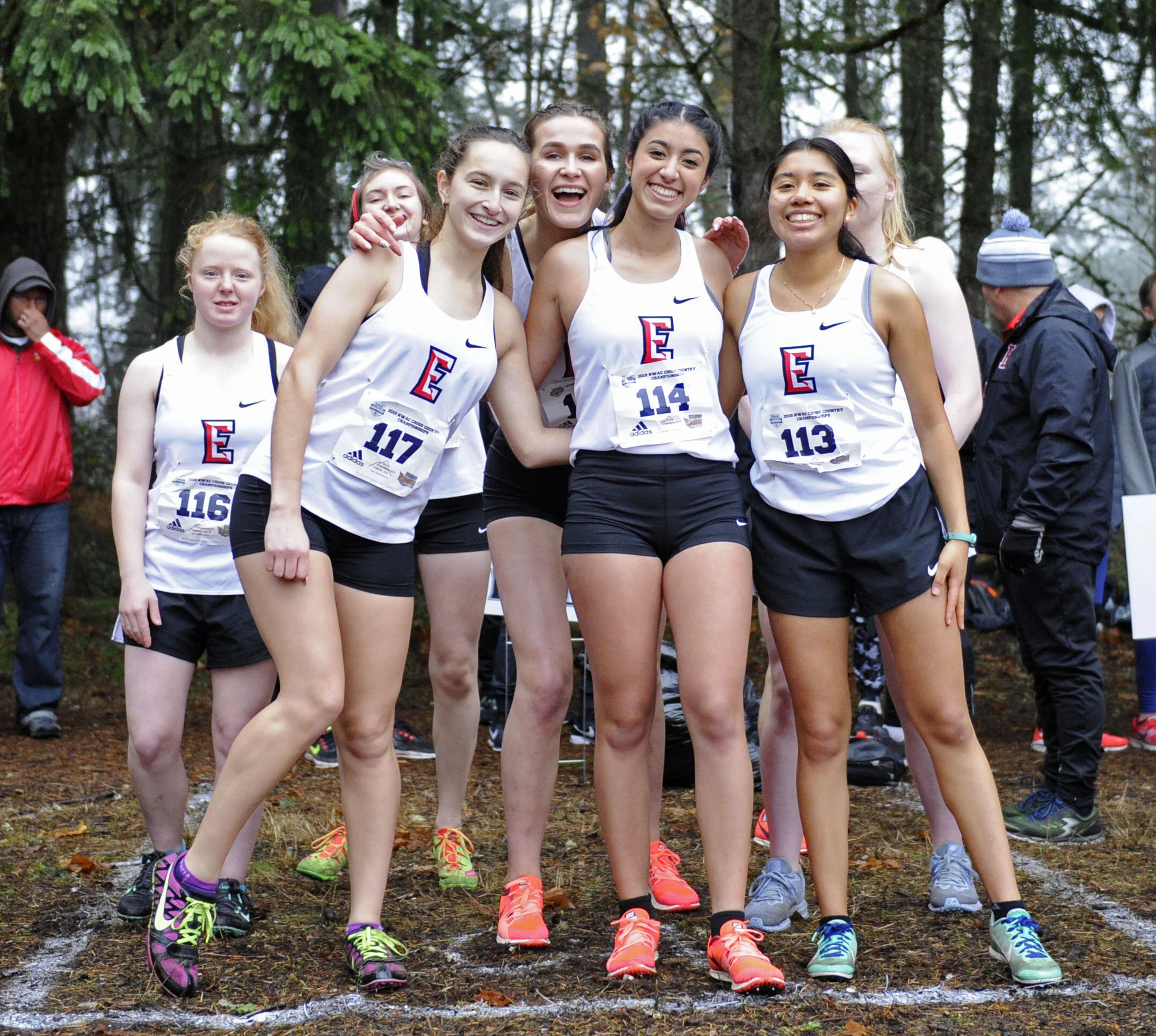 A group of seven young women in athletic running attire posing together outdoors on a wooded trail, smiling for a photo after a race.