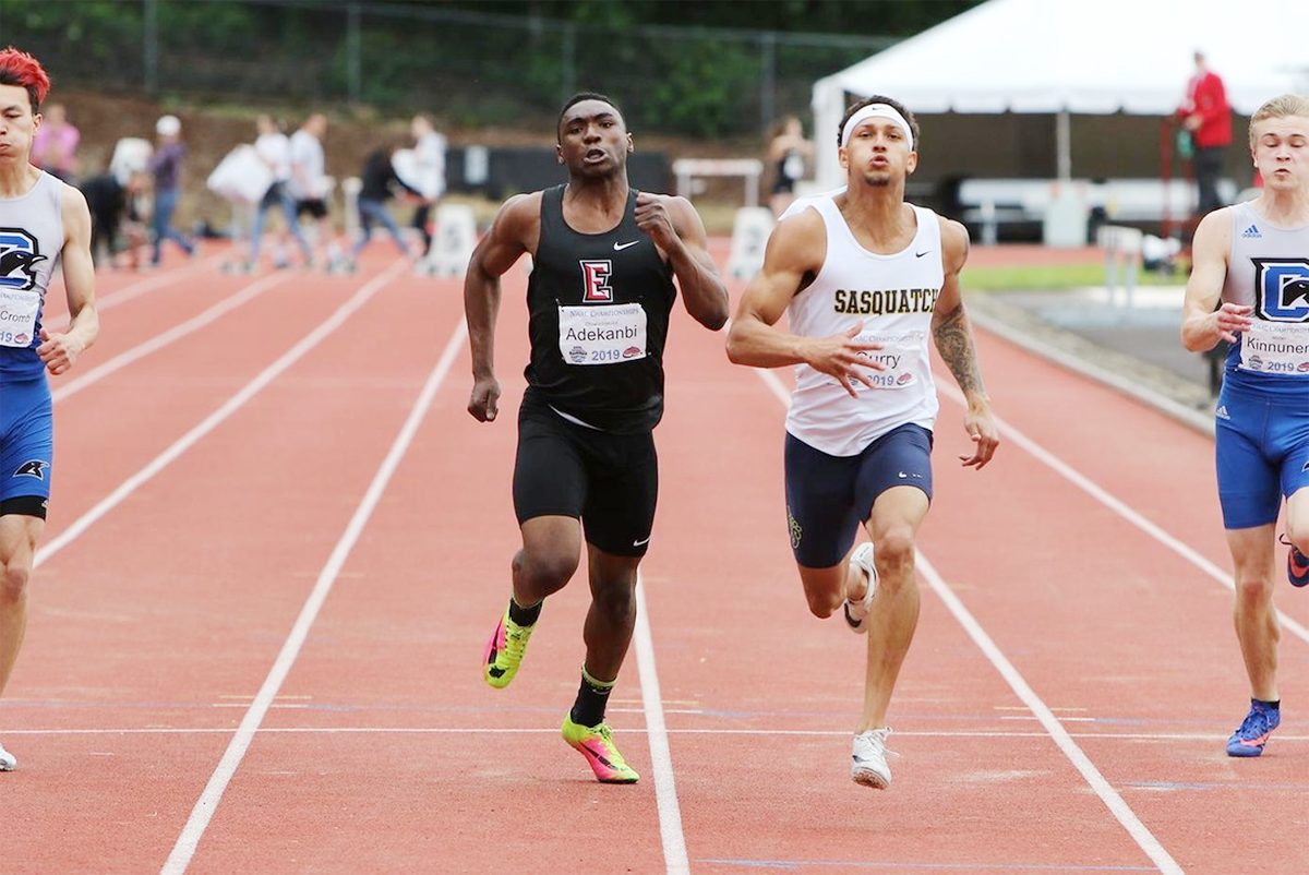 Four male athletes running on a track in a race, with spectators and a tent visible in the background.