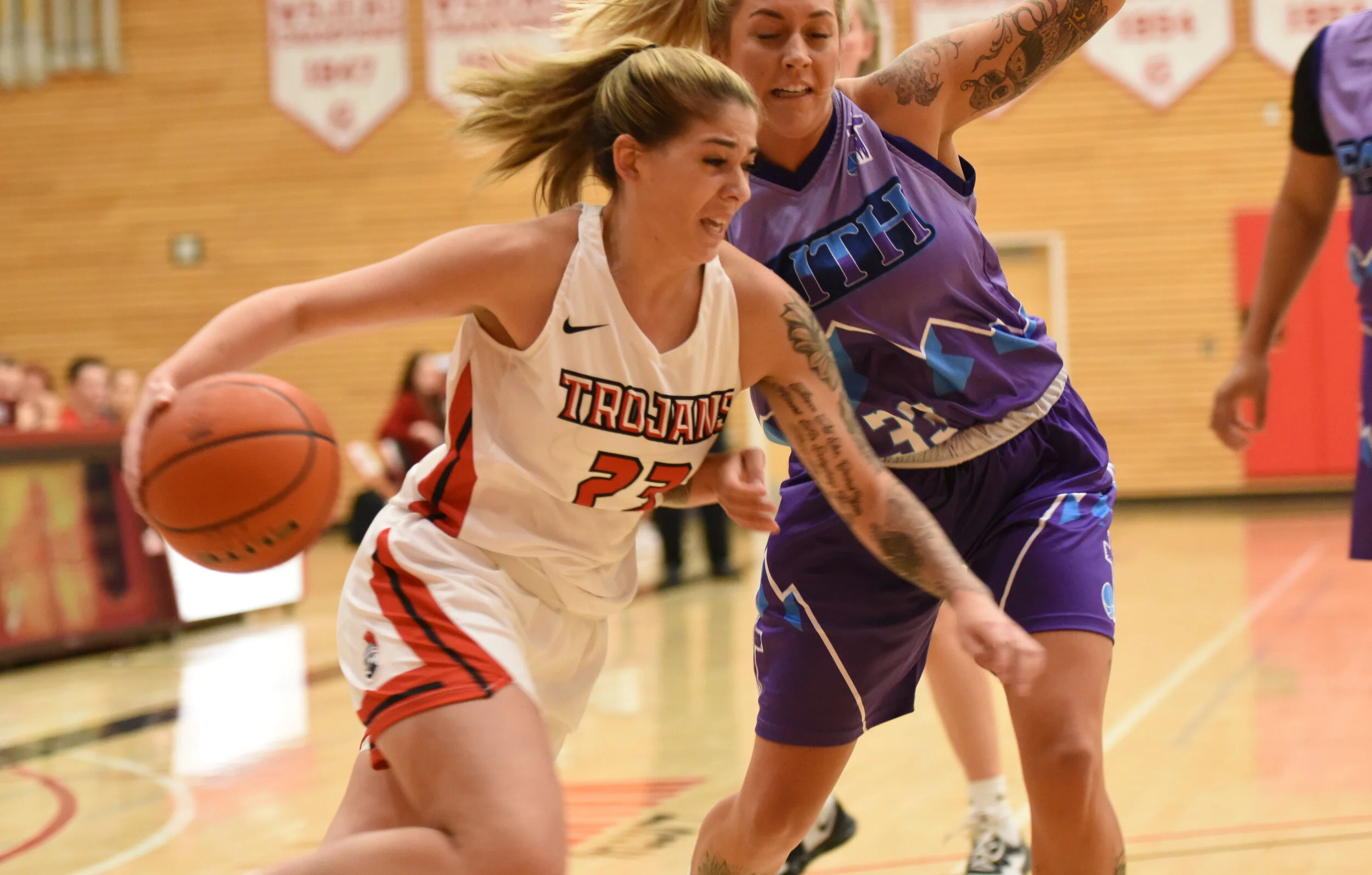 Two female basketball players competing for the ball during a game; one in a white jersey with 'TROJANS' and number 23, the other in a purple jersey with rainbow-colored details.