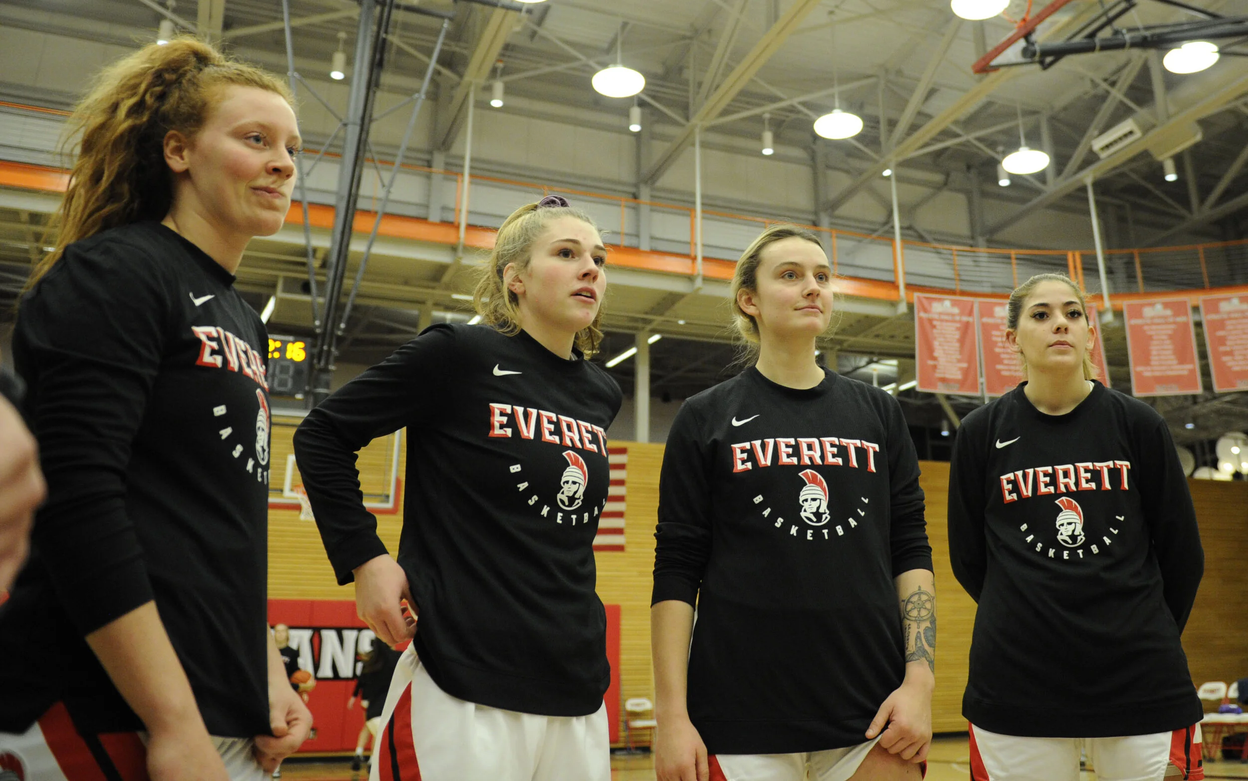 Four female basketball players in black Everett Basketball shirts standing on the court in a gymnasium. They are looking serious, with one having her hand on her hip.