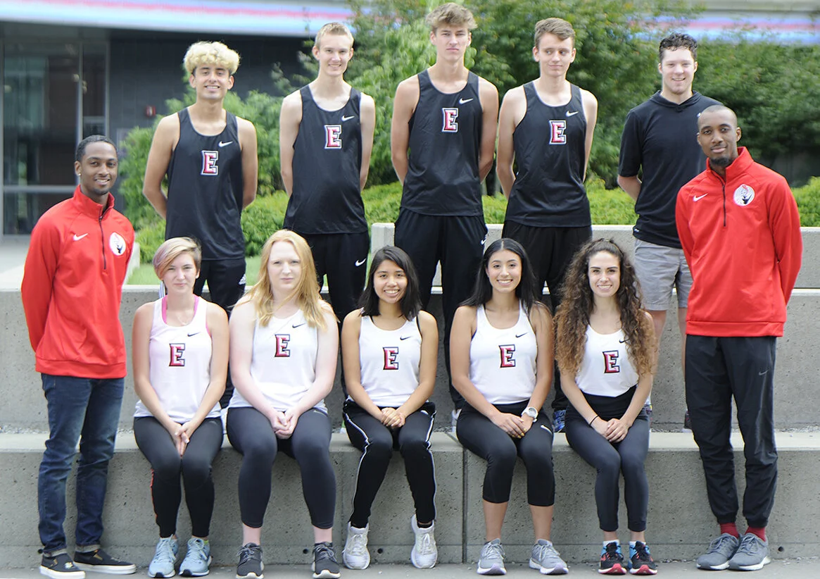 A group of nine young athletes and two coaches posing outdoors on a concrete ledge in front of green bushes and a building. They are wearing athletic clothing with some in black and others in white, all with an 