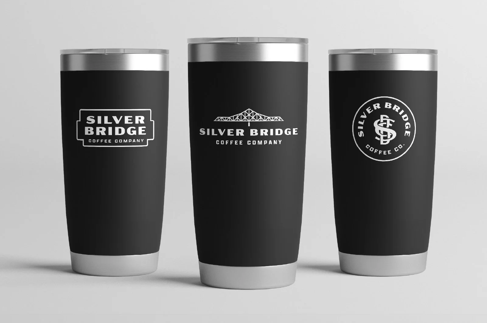 Three black travel mugs with silver lids, each featuring a different Silver Bridge Coffee Co. logo in white.