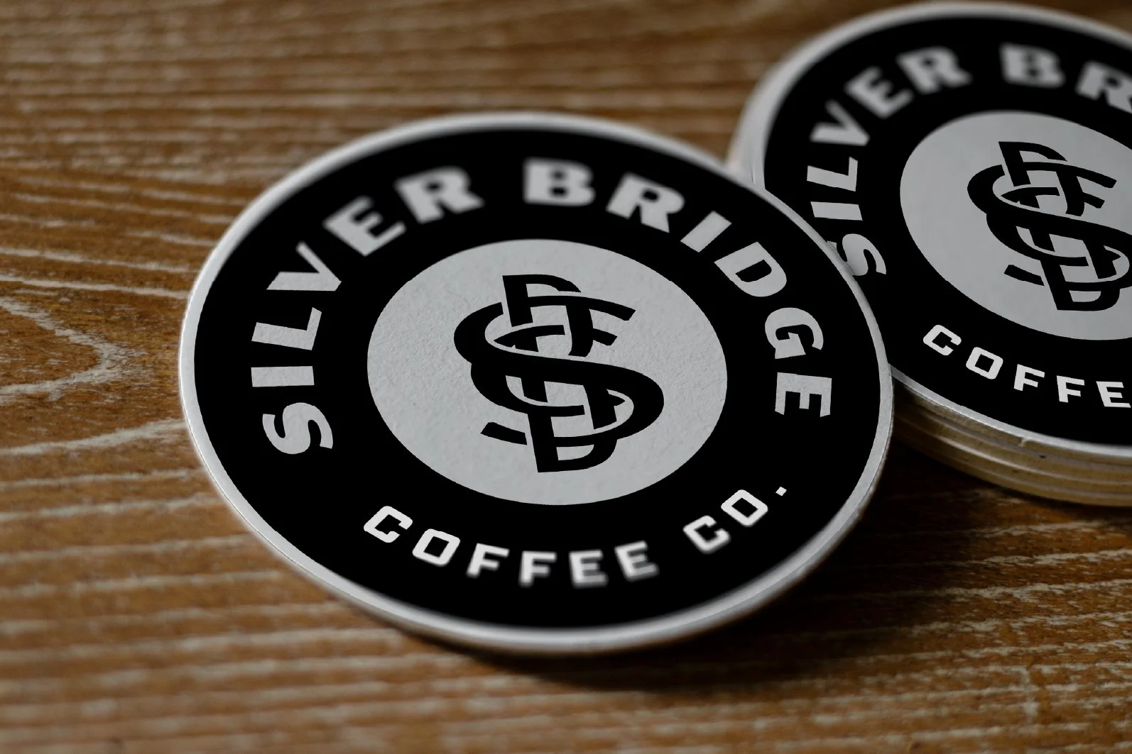 Stacked circular stickers with black and gray design, reading 'Silver Bridoo Coffee Co.' with a stylized intertwined 'S' and 'G' logo in the center, placed on wooden surface.