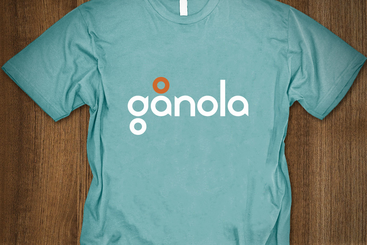 A teal T-shirt with the word "anola" and an abstract logo design printed on the front, laid flat on a wooden surface.