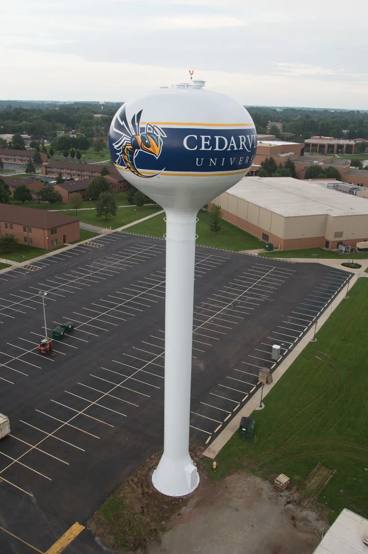 A tall white water tower with a university logo and the words 'Cedarville University' painted on it, situated above an empty parking lot.