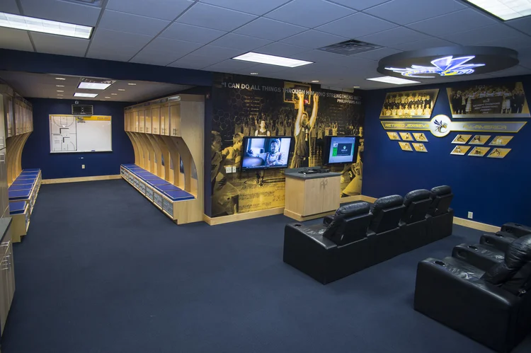 A sports team locker room with wooden lockers and blue benches, large flat-screen TVs, motivational wall art, and black leather chairs for viewing.