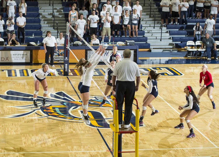 A volleyball game in progress on a gymnasium court with players jumping at the net and others preparing to receive the ball, spectators in the stands.