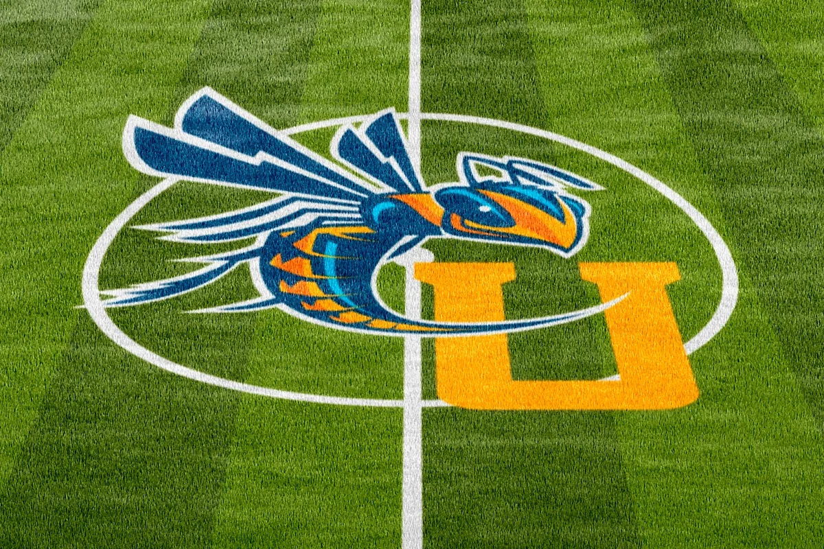 Logo of the UConn Huskies football team on a green field, featuring a stylized blue and orange hawk and a large yellow "U".