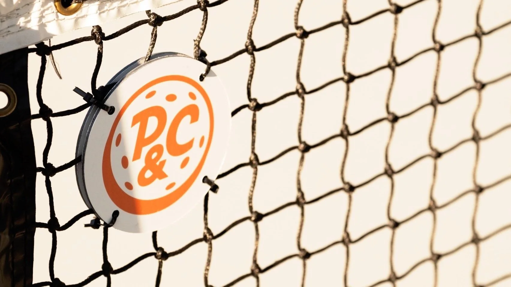 Close-up of a sign with the text 'P.C.' in orange, attached to a black netted fence.