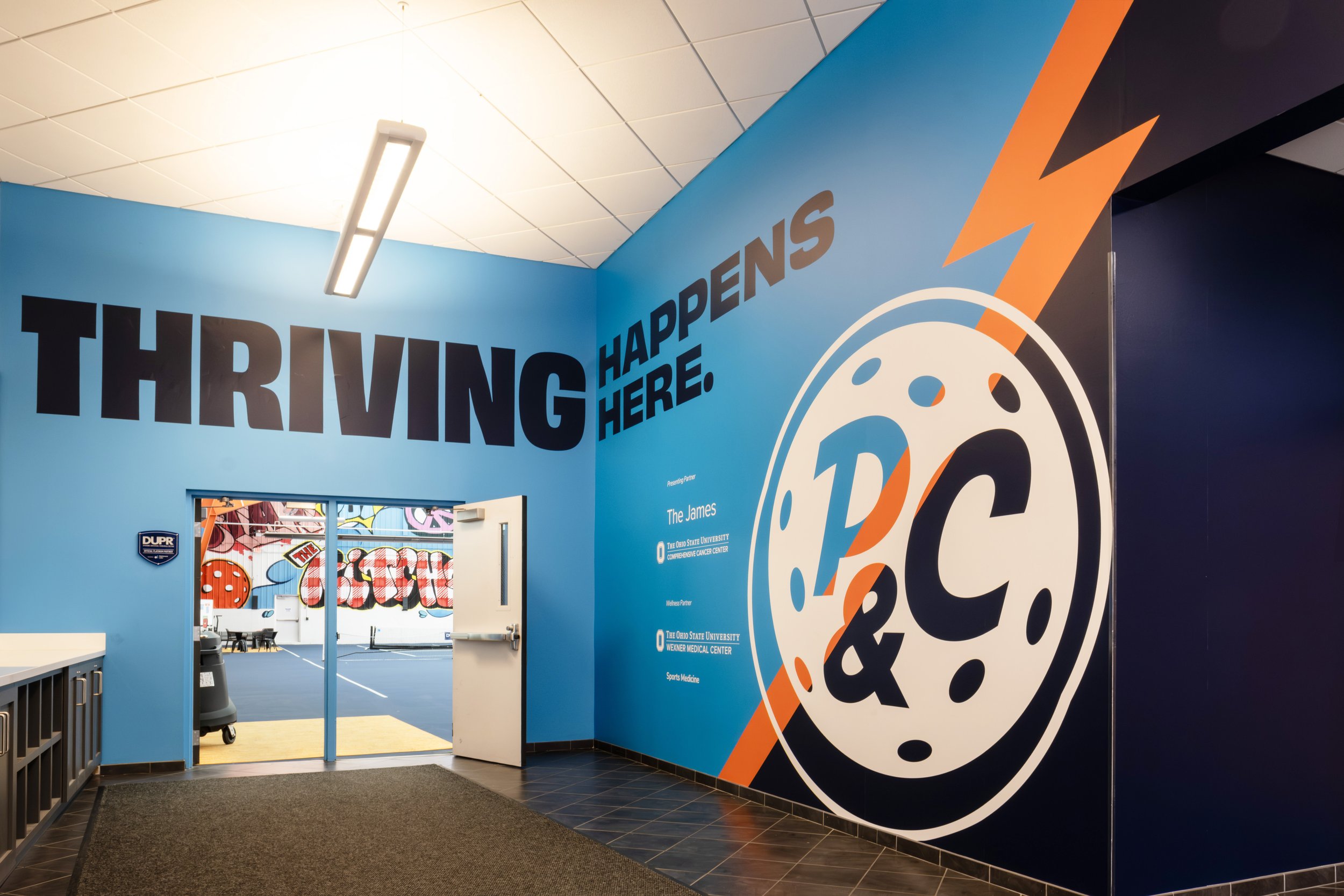 A colorful indoor space with a blue wall featuring large text that reads 'THRIVING HAPPENS HERE.' Next to the text, there is a large clock graphic with 'P & C' inside, accented by an orange lightning bolt. An open door reveals a gym or sports area wi