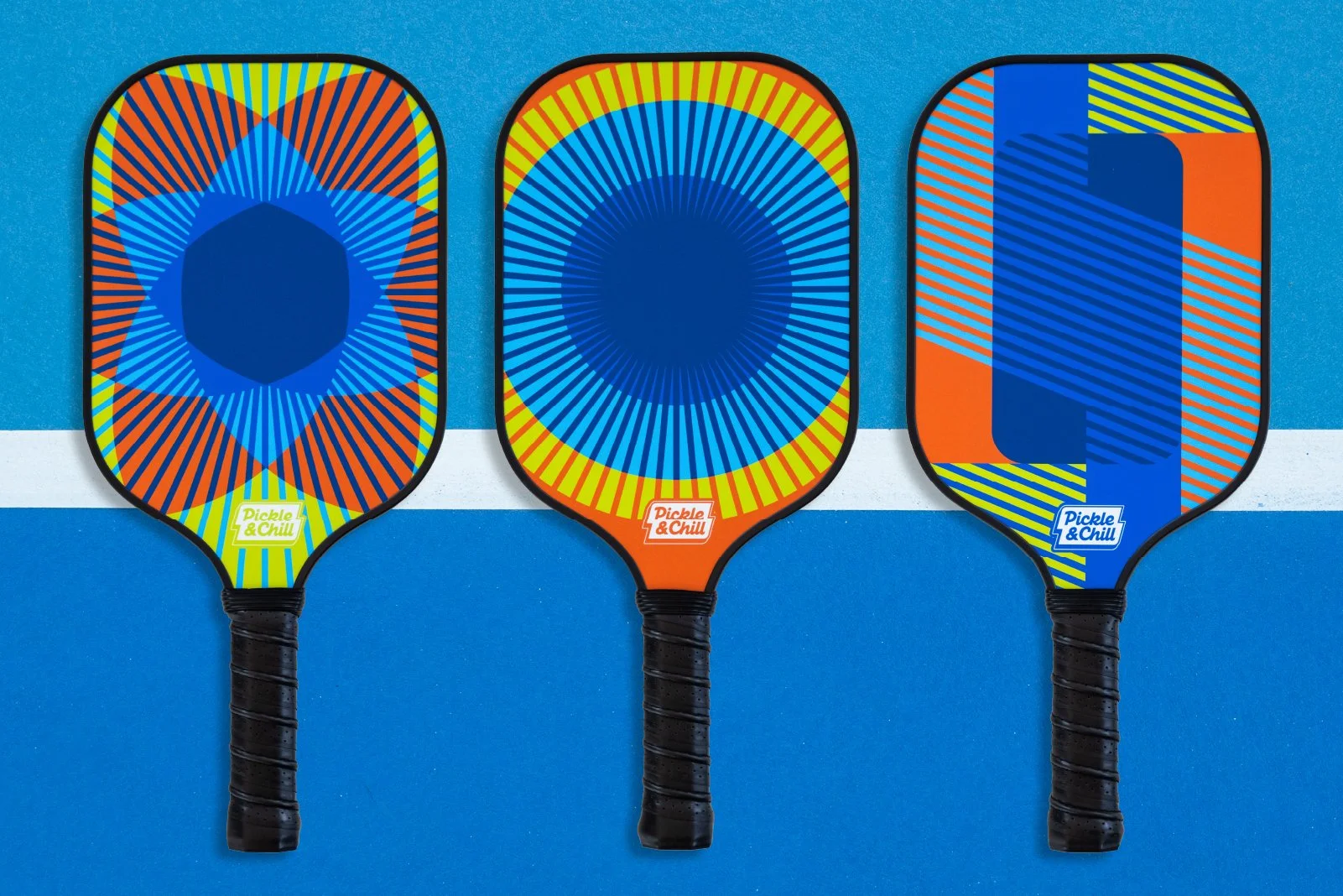 Three colorful pickleball paddles with geometric patterns, arranged side by side on a blue and white court surface.