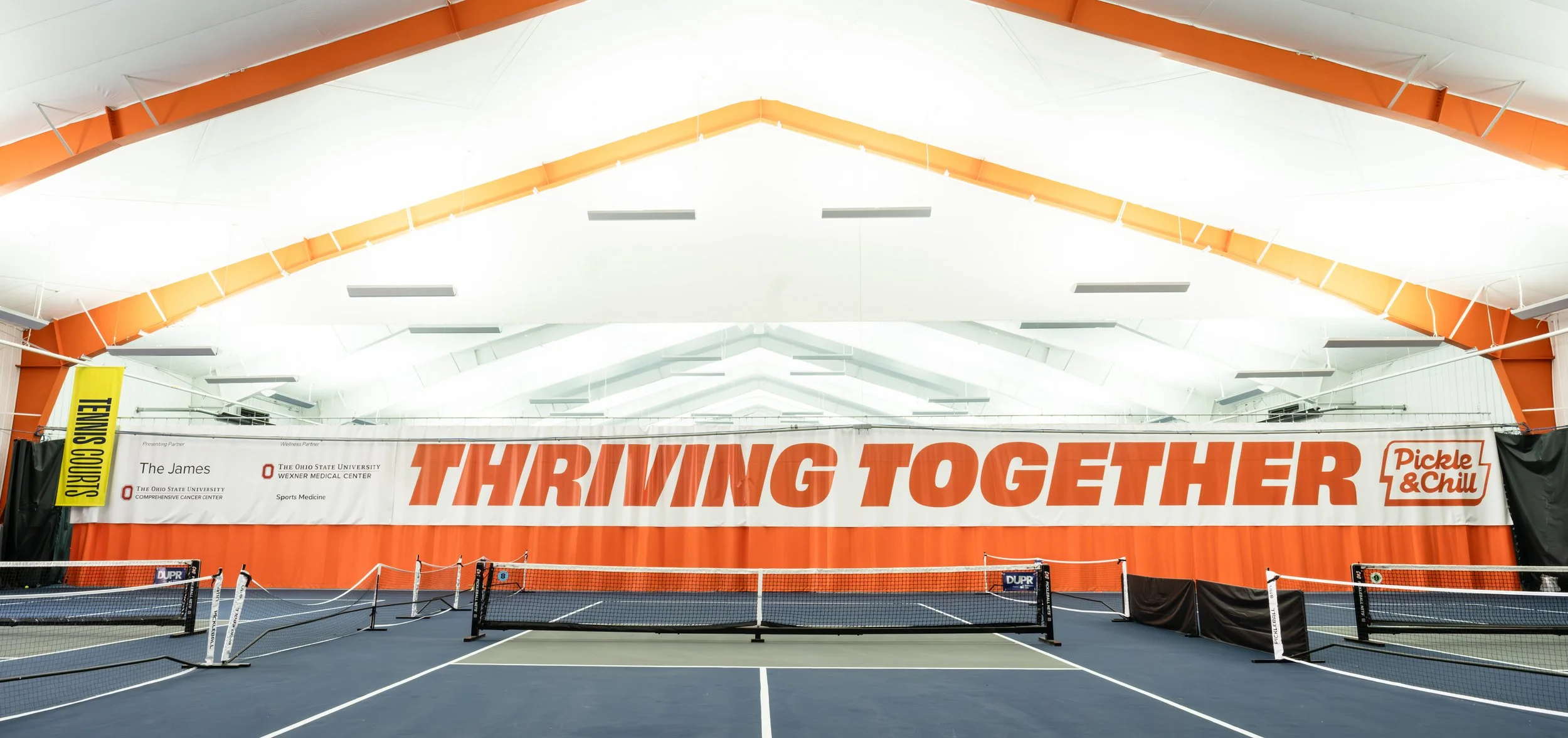 Indoor tennis court with a large banner reading 'Thriving Together' and a Pickle & Chill logo, surrounded by black curtains and nets.