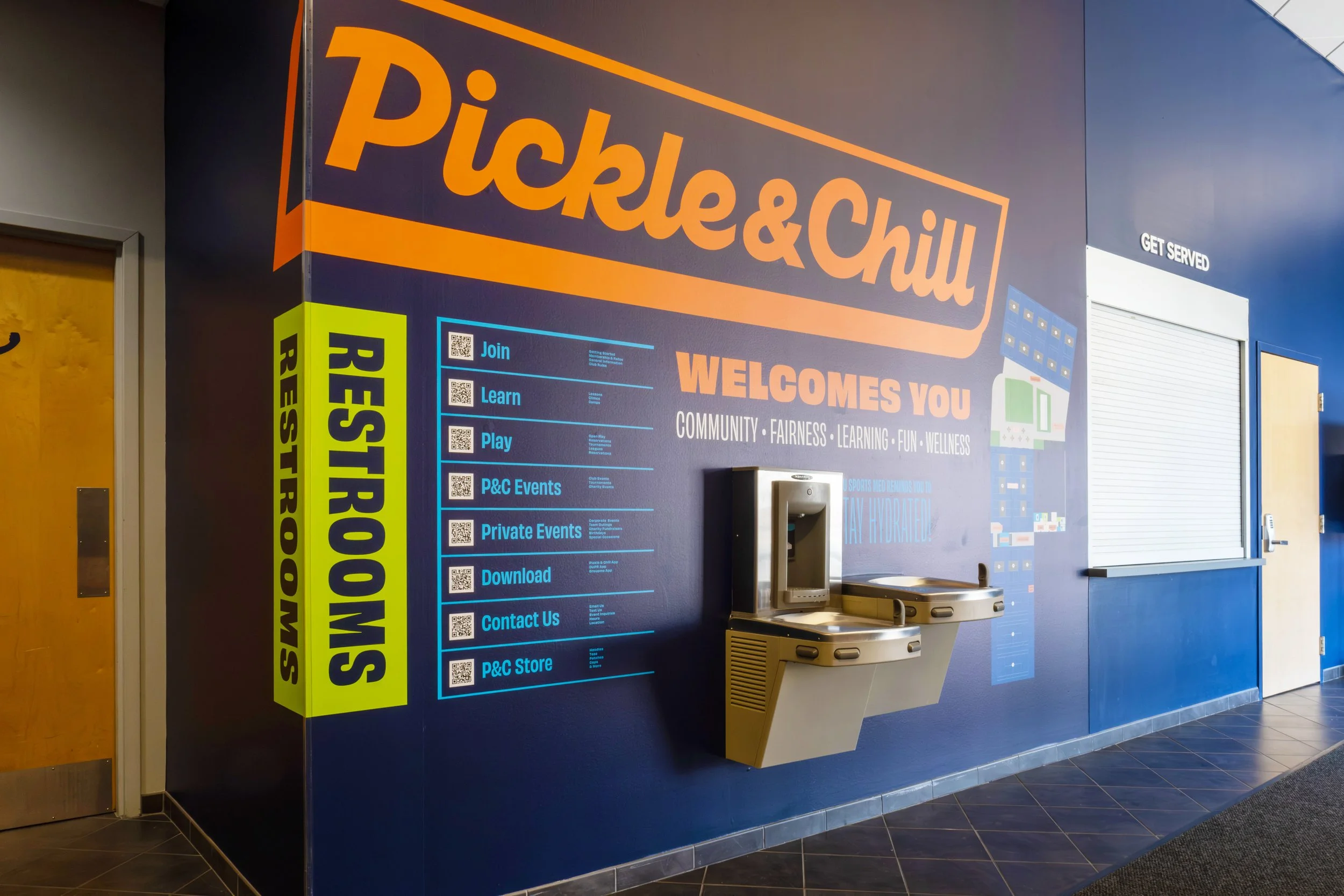 Wall at Pickle & Chill with colorful signage and information, including a menu, event info, and a water fountain, with a closed service window.