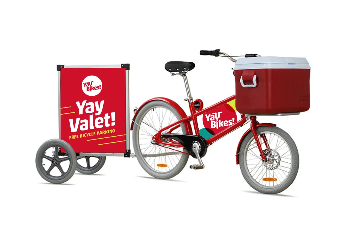 Red bicycle with a large red basket attached to the front and a sign on a trailer attached to the back indicating free bicycle parking with the text 'Yay Valet! FREE BICYCLE PARKING'