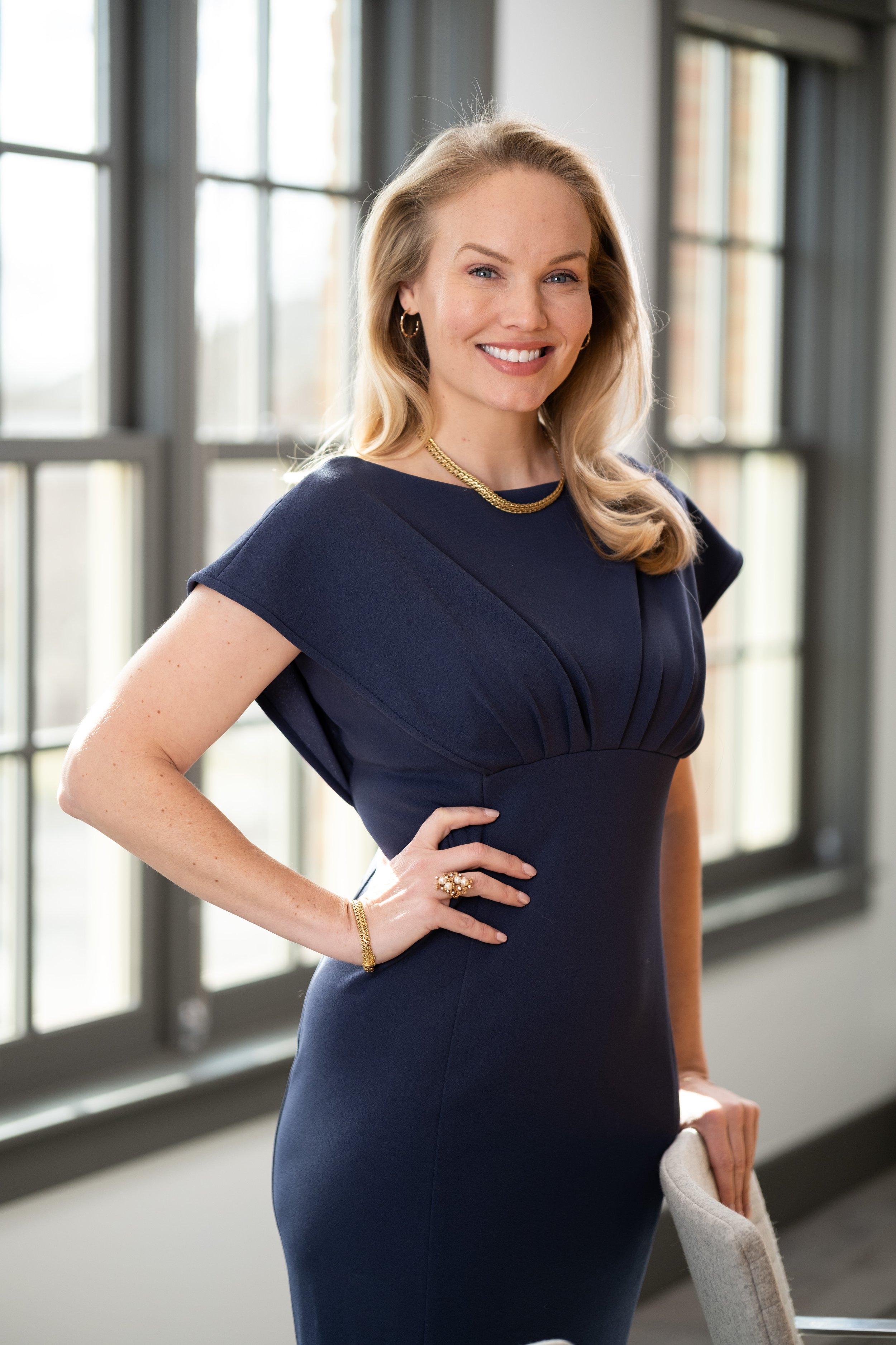 A woman with blonde hair, wearing a navy blue dress, standing indoors near large windows, smiling, with her left hand on her hip.