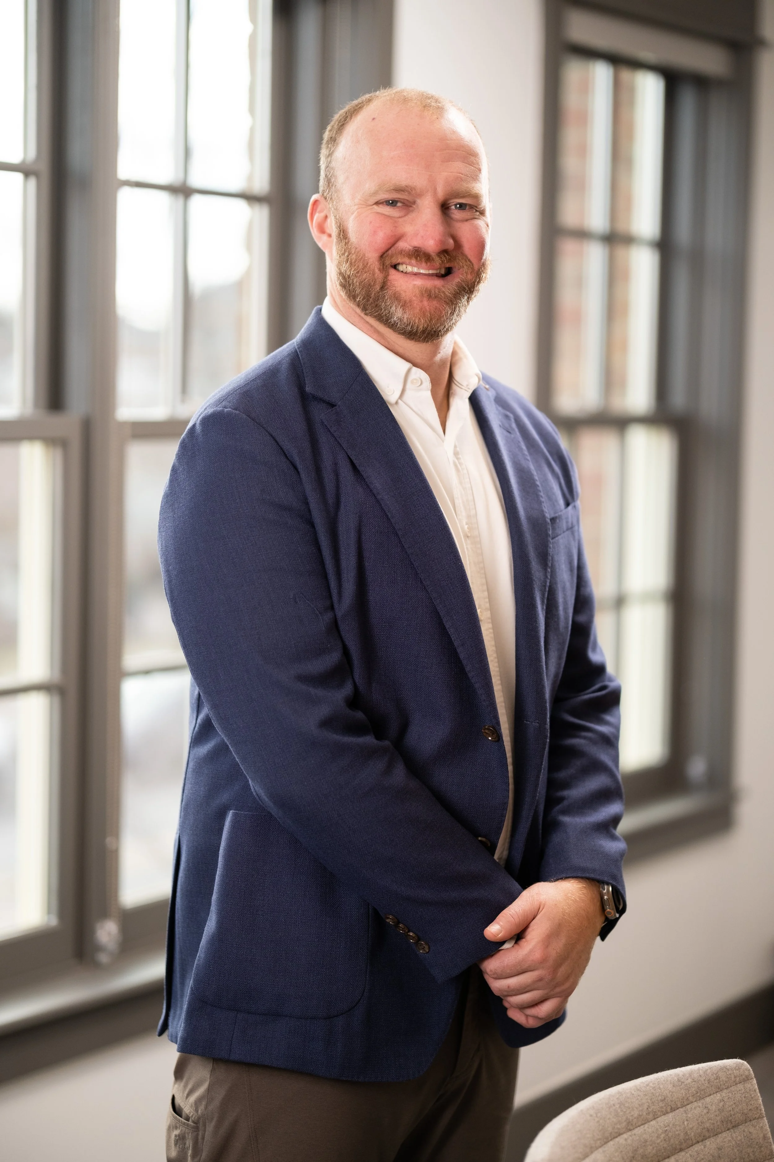 A man with a beard, wearing a navy blazer and white shirt, standing indoors in front of large windows, smiling at the camera.