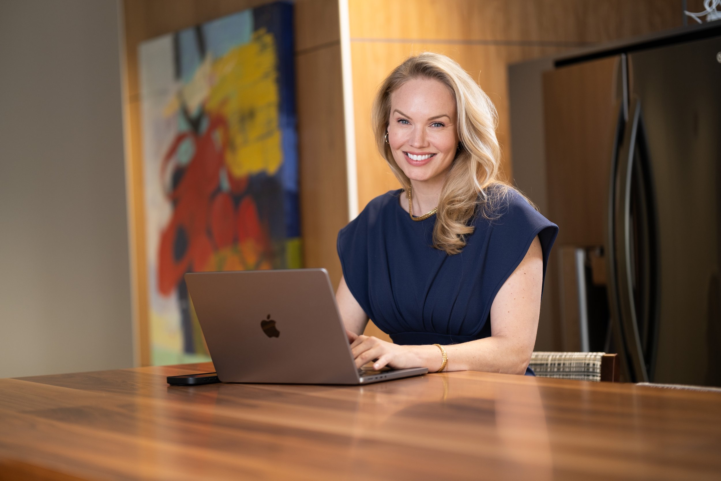 A woman with blonde hair wearing a navy blue dress sitting at a wooden table, smiling, with a laptop open in front of her and abstract colorful artwork on the wall behind her.