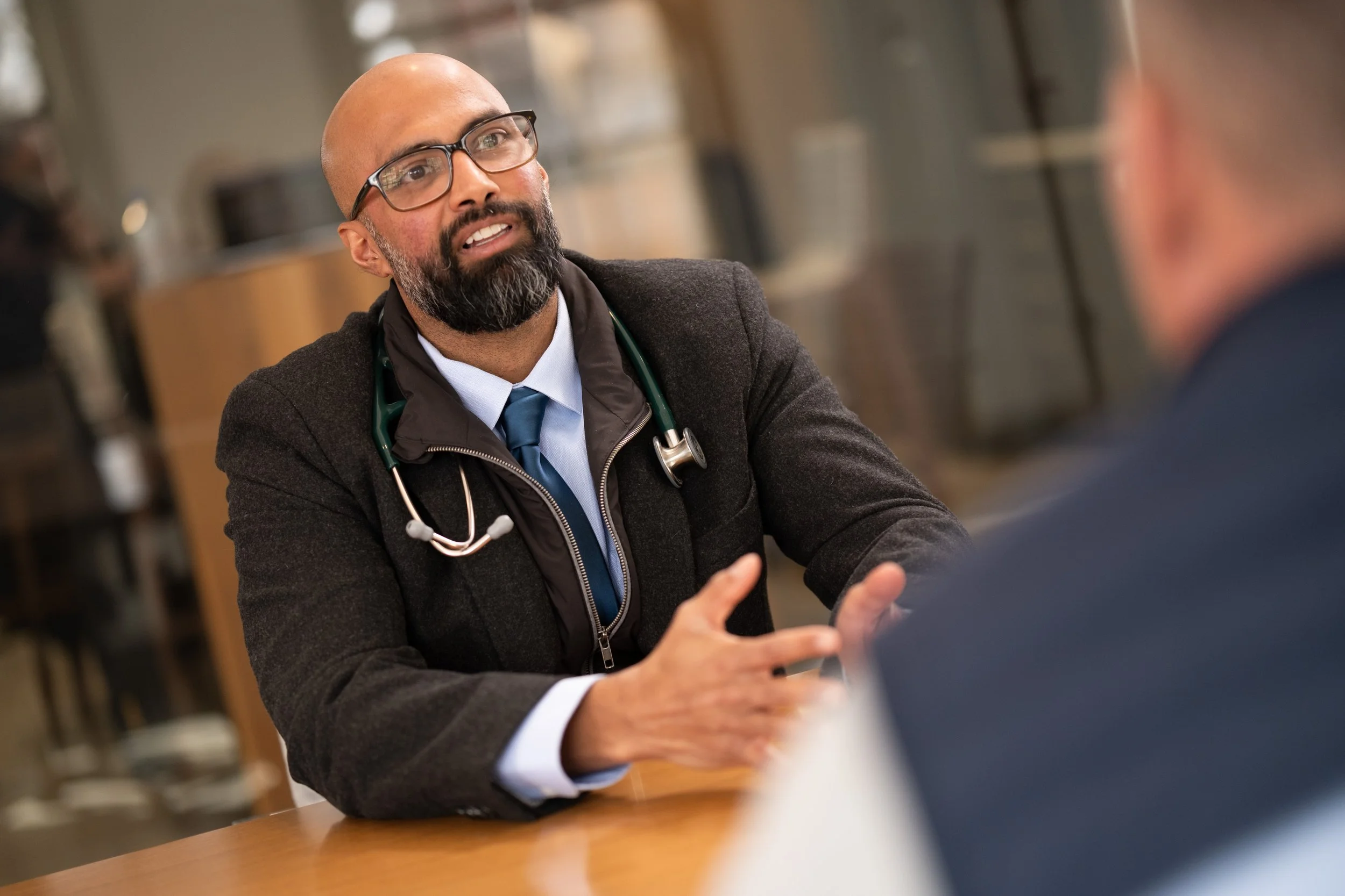 A man wearing glasses, a dark coat, and a stethoscope around his neck is engaged in a conversation at a table with another person. The setting appears to be an indoor area, possibly a medical or professional environment.