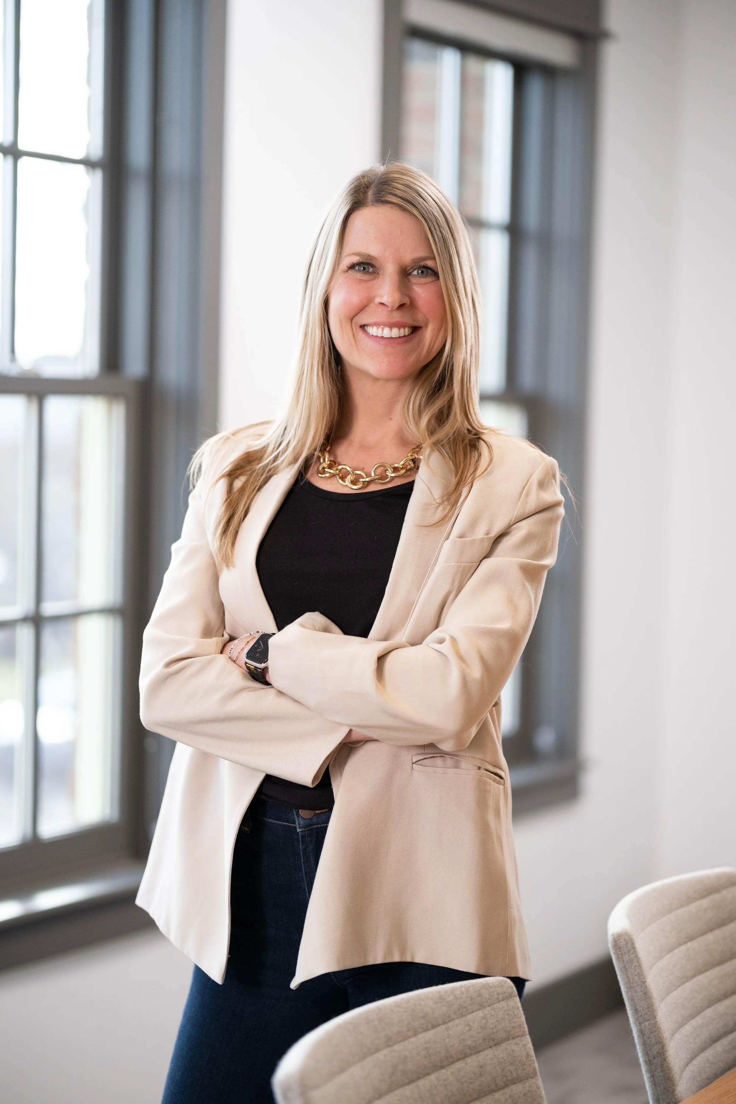 A smiling woman with long blonde hair wearing a beige blazer, black shirt, and gold chain necklace in a bright office setting.
