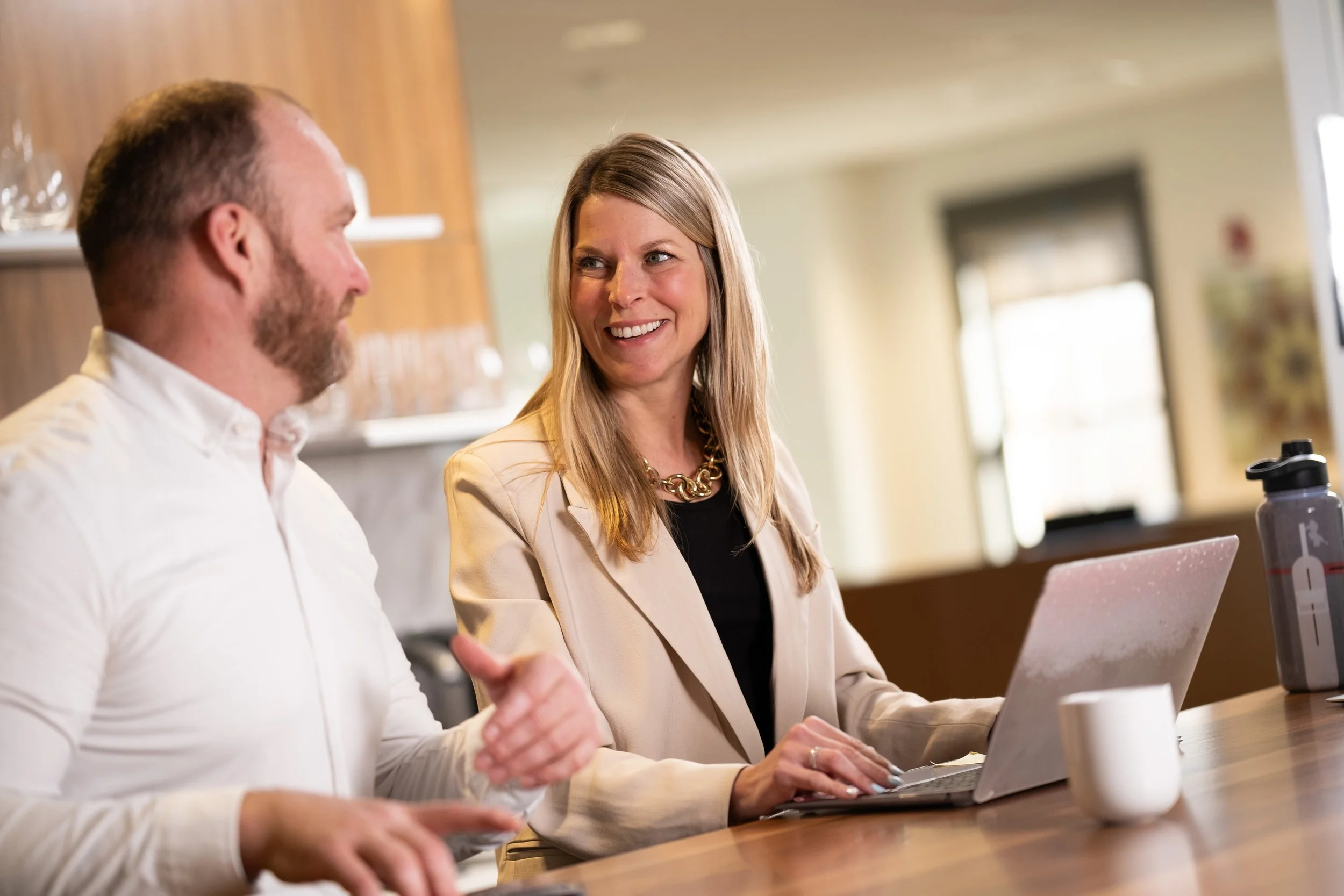 A woman smiling and talking with a man in a casual work or meeting space, with a laptop, a water bottle, and a mug on the table.