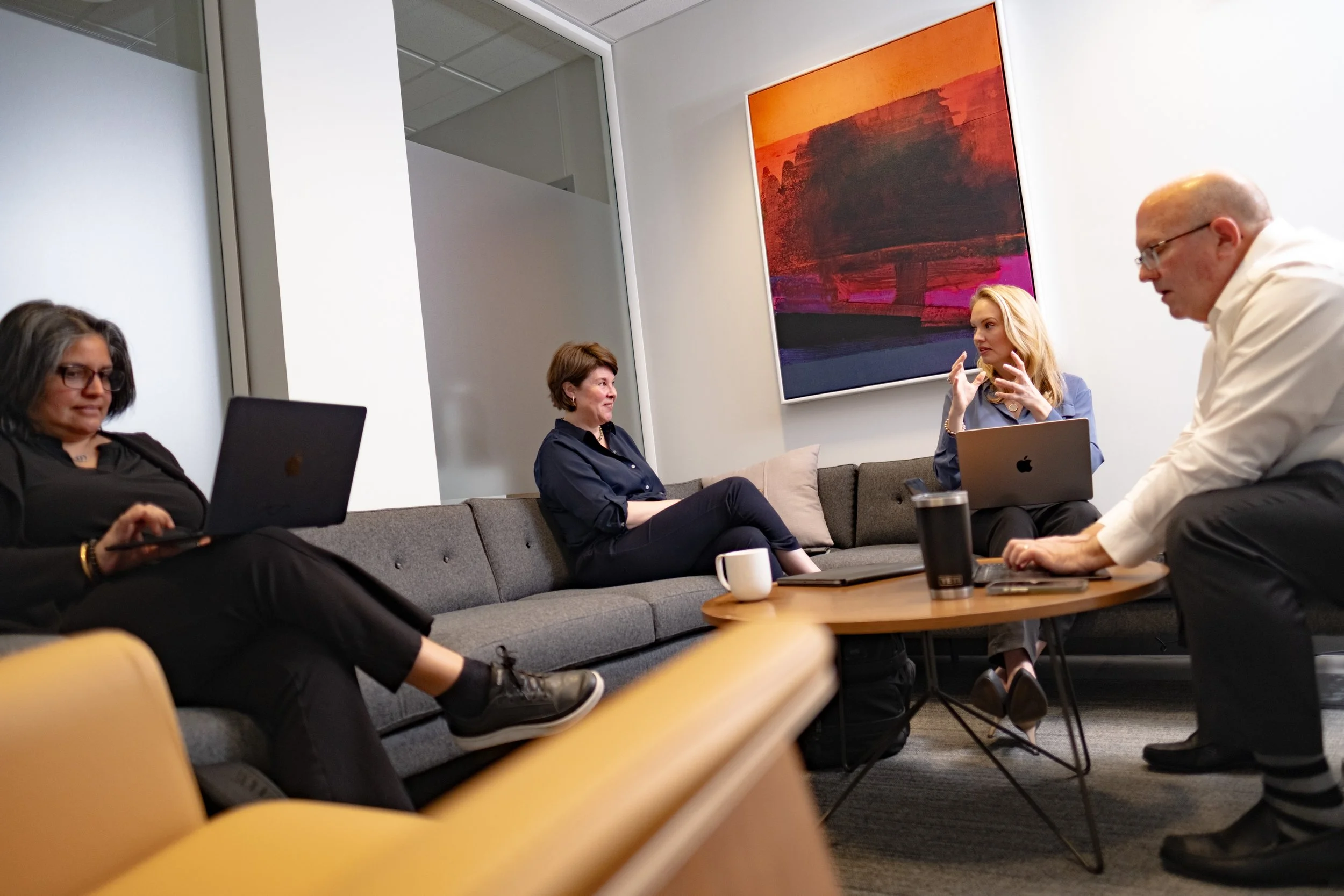 Four professionals in a meeting room, two women and one man working on laptops, engaging in a discussion, with a colorful abstract painting on the wall.