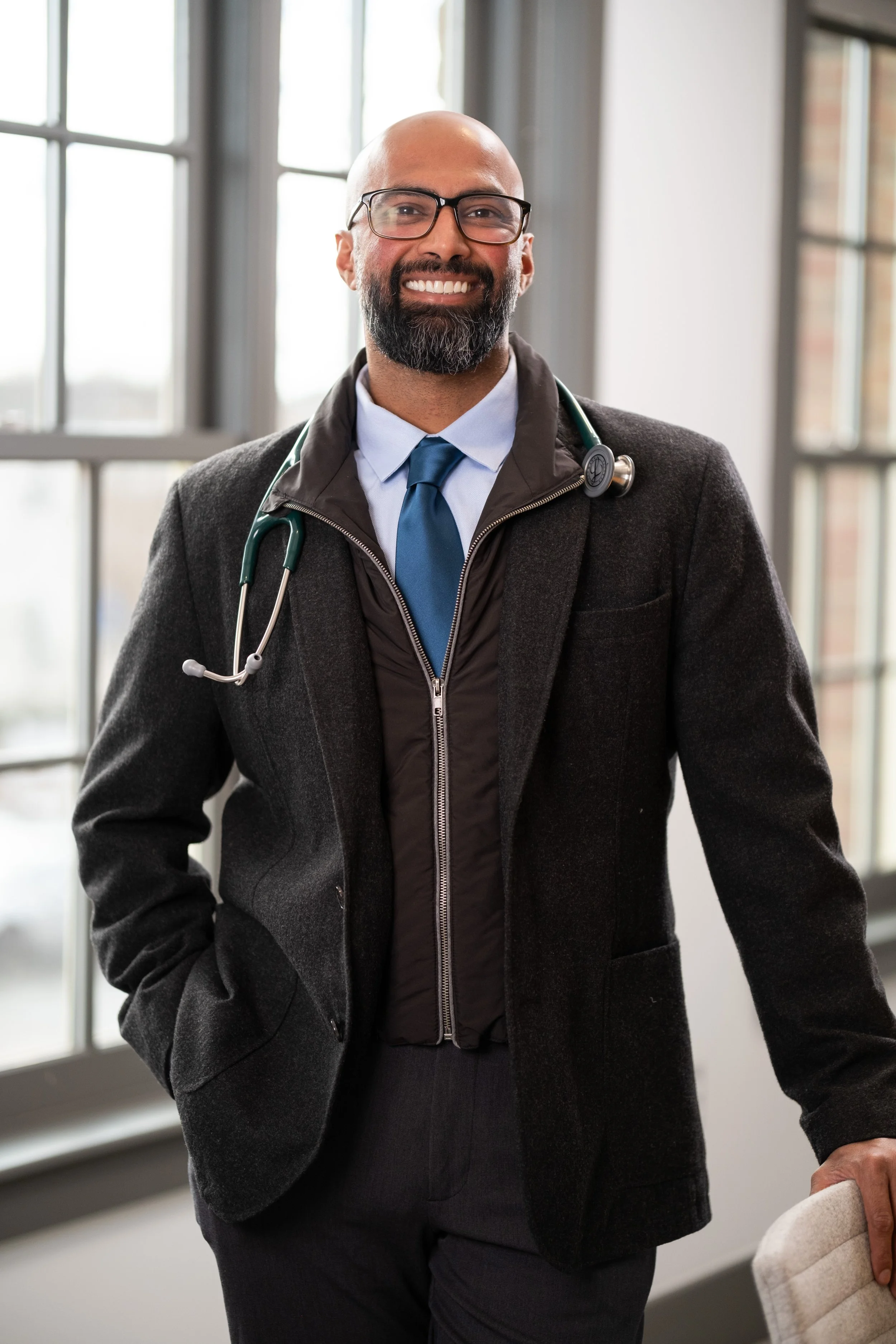 A smiling male doctor with a beard and glasses, wearing a blue dress shirt, tie, and black coat, with a stethoscope around his neck, standing in a bright room with large windows.
