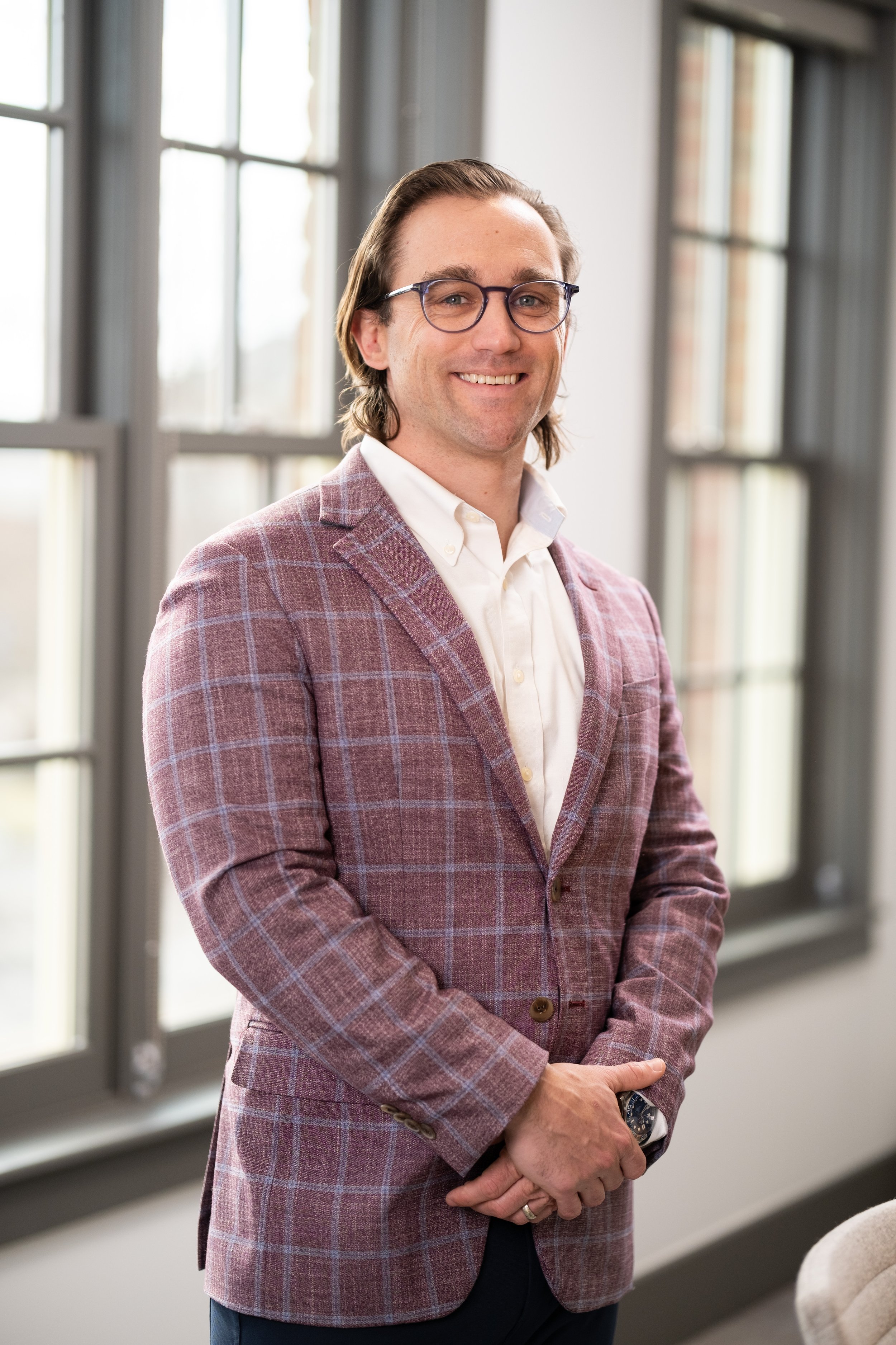 A man with glasses in a plaid blazer and white shirt smiling, standing in front of large windows in a well-lit room.