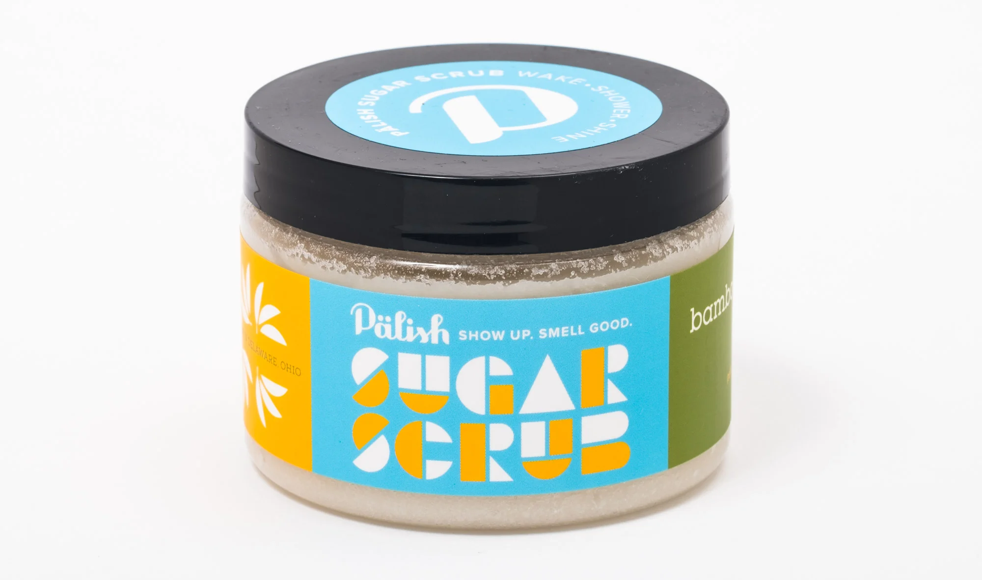A jar of sugar scrub with a black lid and colorful label featuring blue, yellow, and green sections, and the words "Sugar Scrub" in bold, stylized letters.