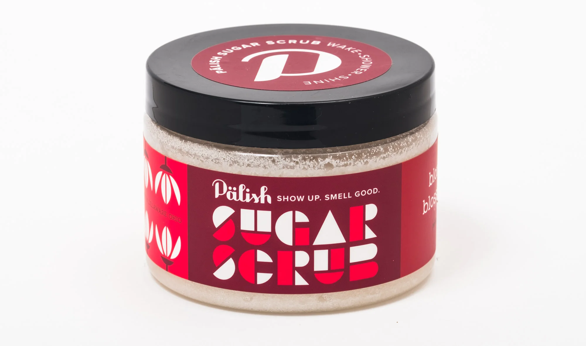 A jar of Pālish sugar scrub with a black lid and red label featuring white and red geometric text and designs, placed against a white background.